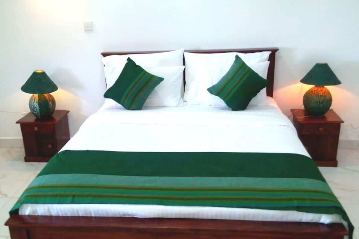 Una cama bien hecha con una colcha verde y blanca a rayas, dos almohadas verdes y dos mesitas de noche de madera con lámparas.