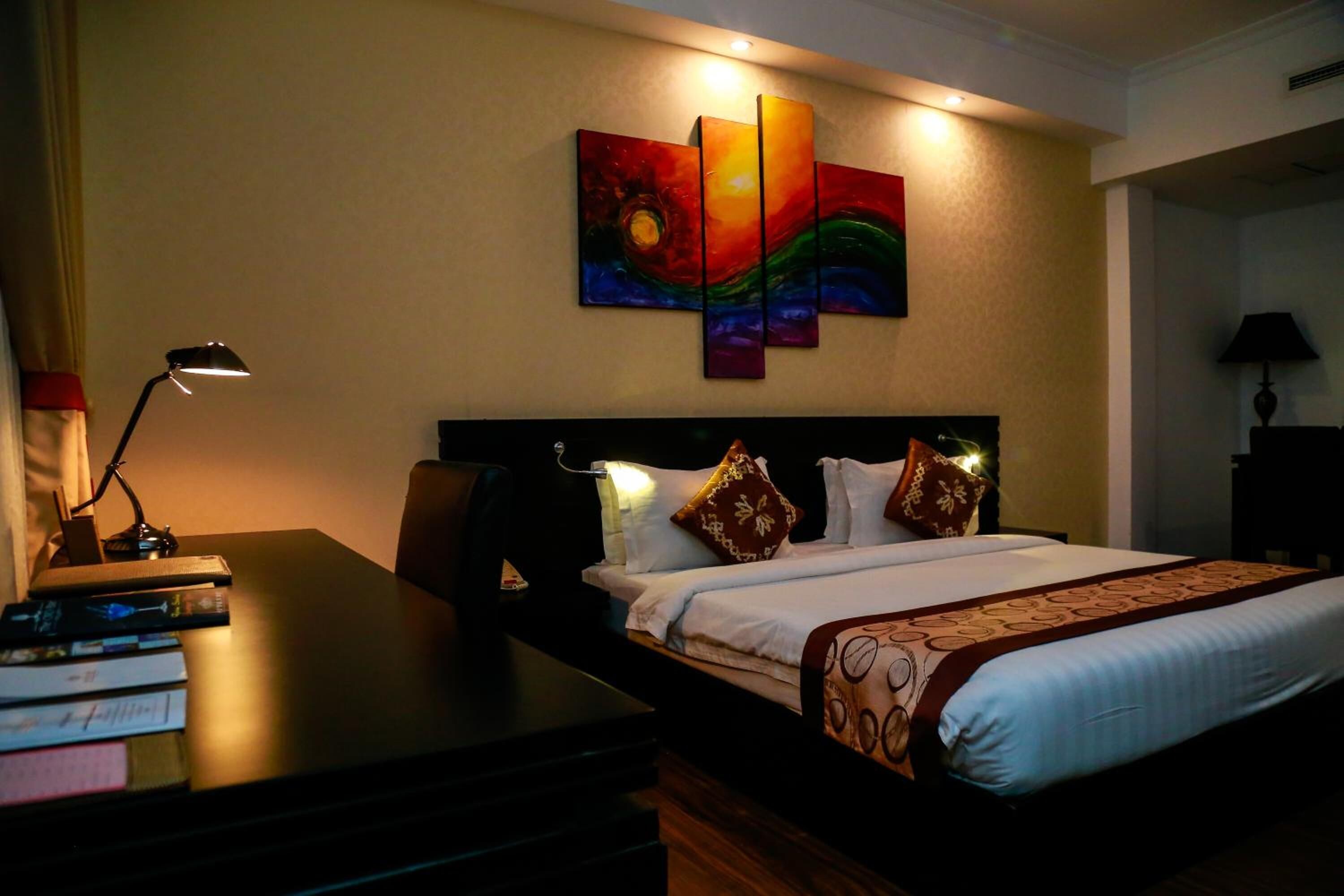 Una habitación de hotel con una cama, un escritorio con libros, una lámpara y un cuadro en la pared.