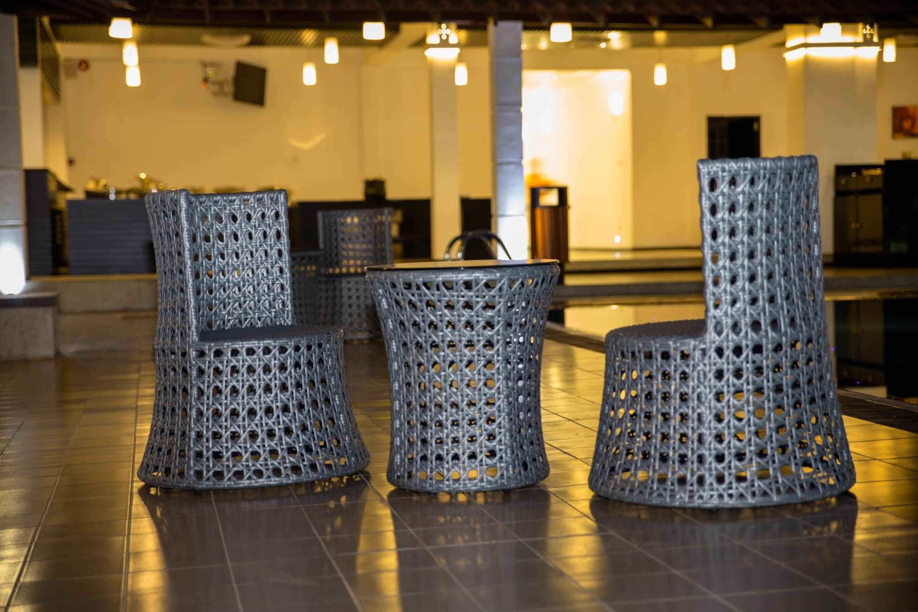 Uma área de estar com três cadeiras de vime e uma mesa pequena, dispostas sobre um piso de azulejos em um espaço interno bem iluminado.