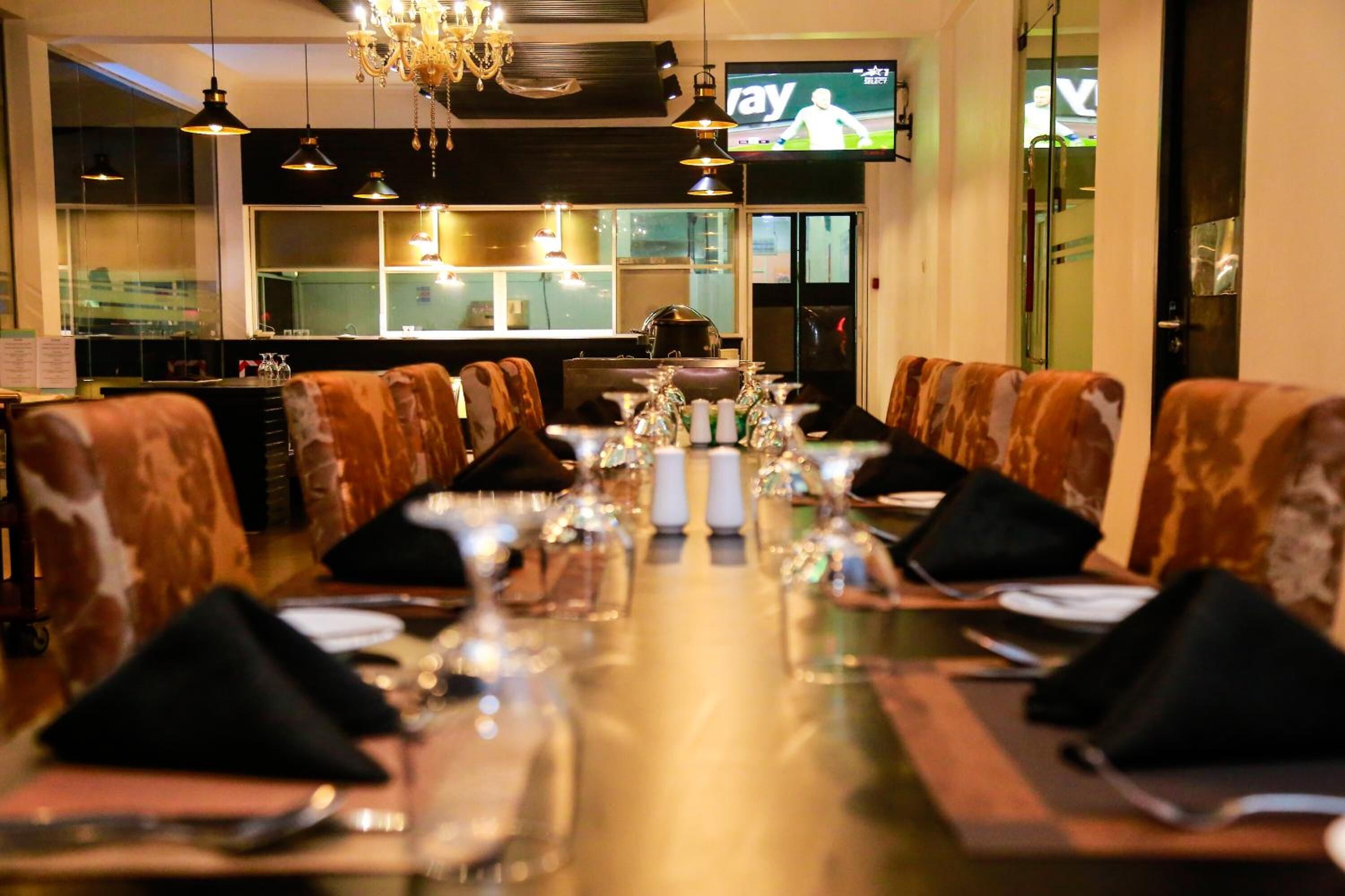 A restaurant with a long table set for a meal, a television screen in the background, and a view of the kitchen through the window.