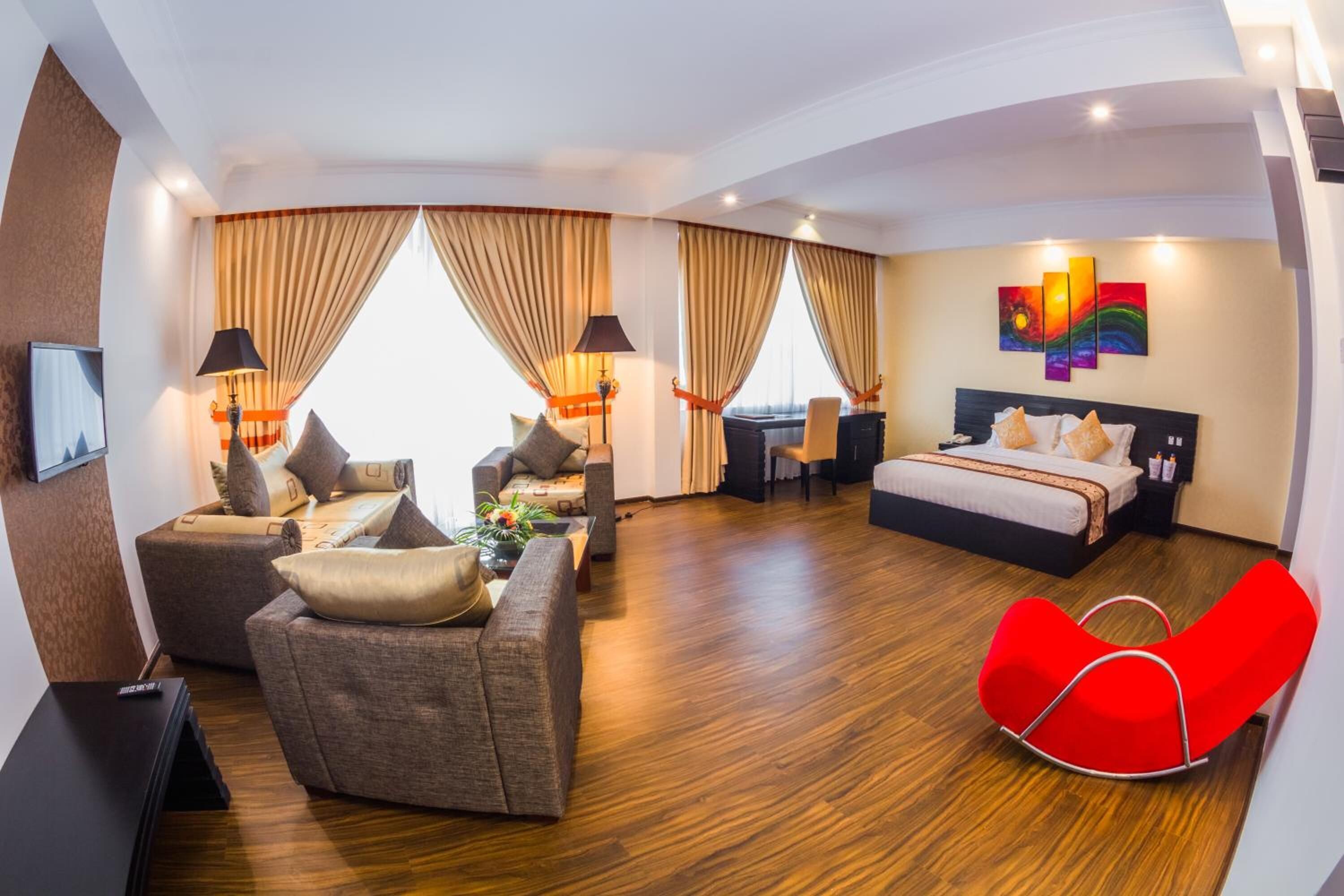 A modern hotel room with a sofa, a red chair, a bed, a desk, and a TV.