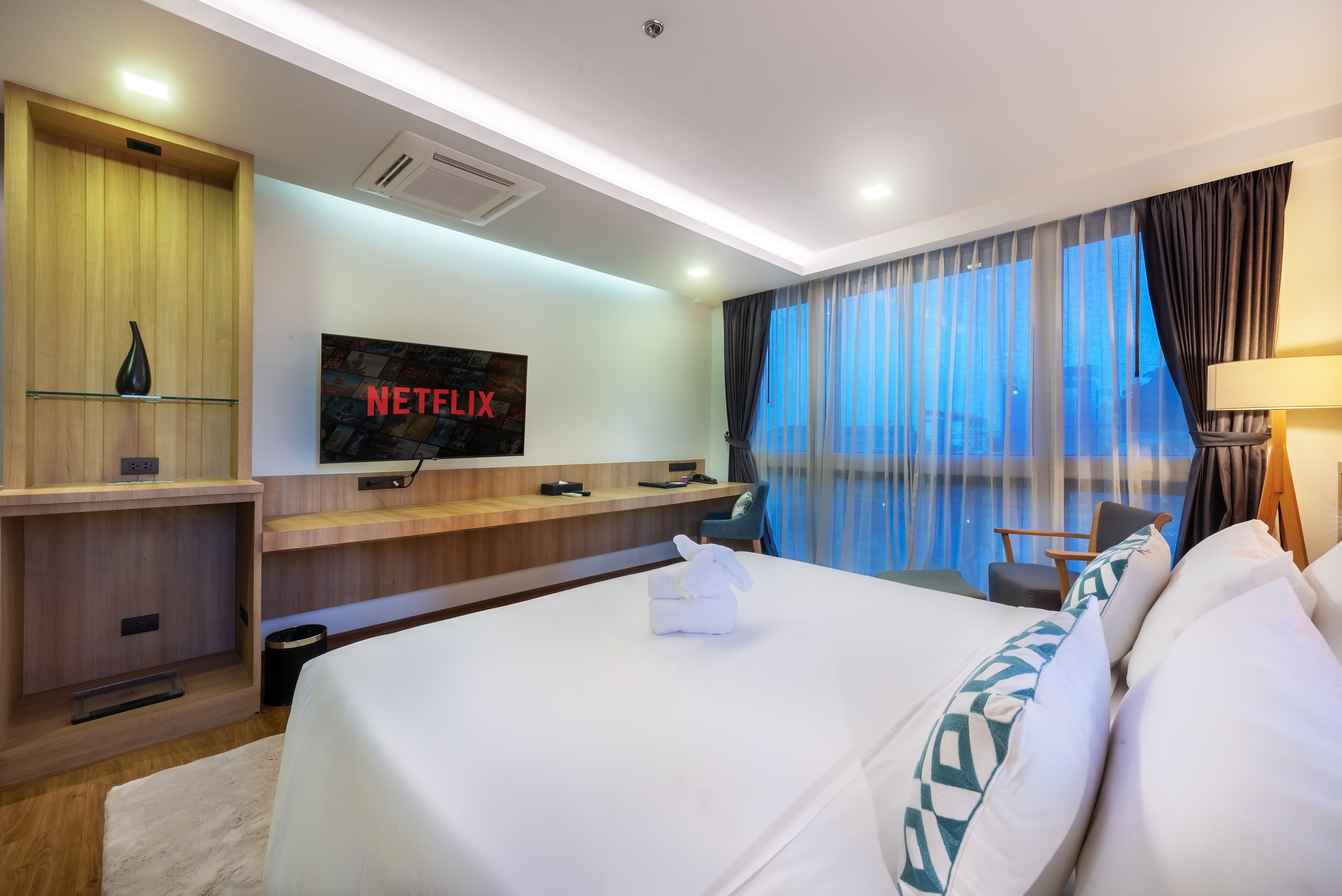 A hotel room with a large bed, a TV mounted on the wall, a wooden desk, and a window with curtains.