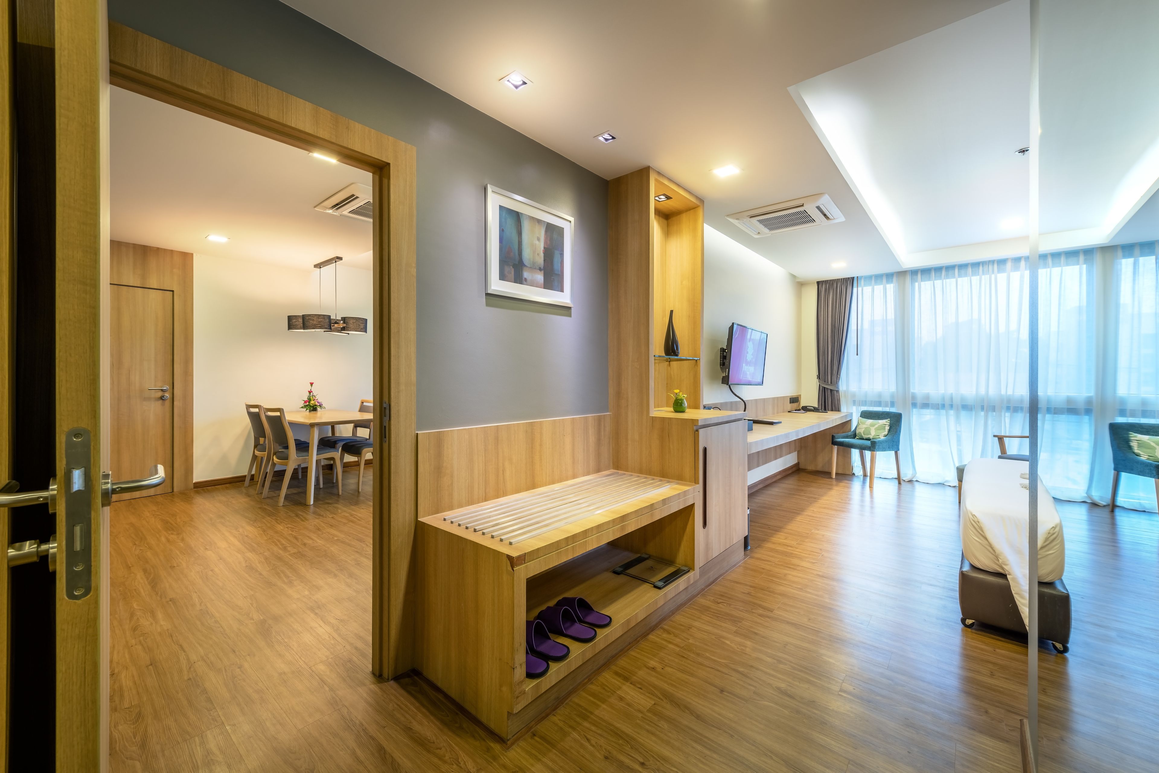 A modern hotel room with a wooden bench, a dining area, a TV, and a large window with curtains.