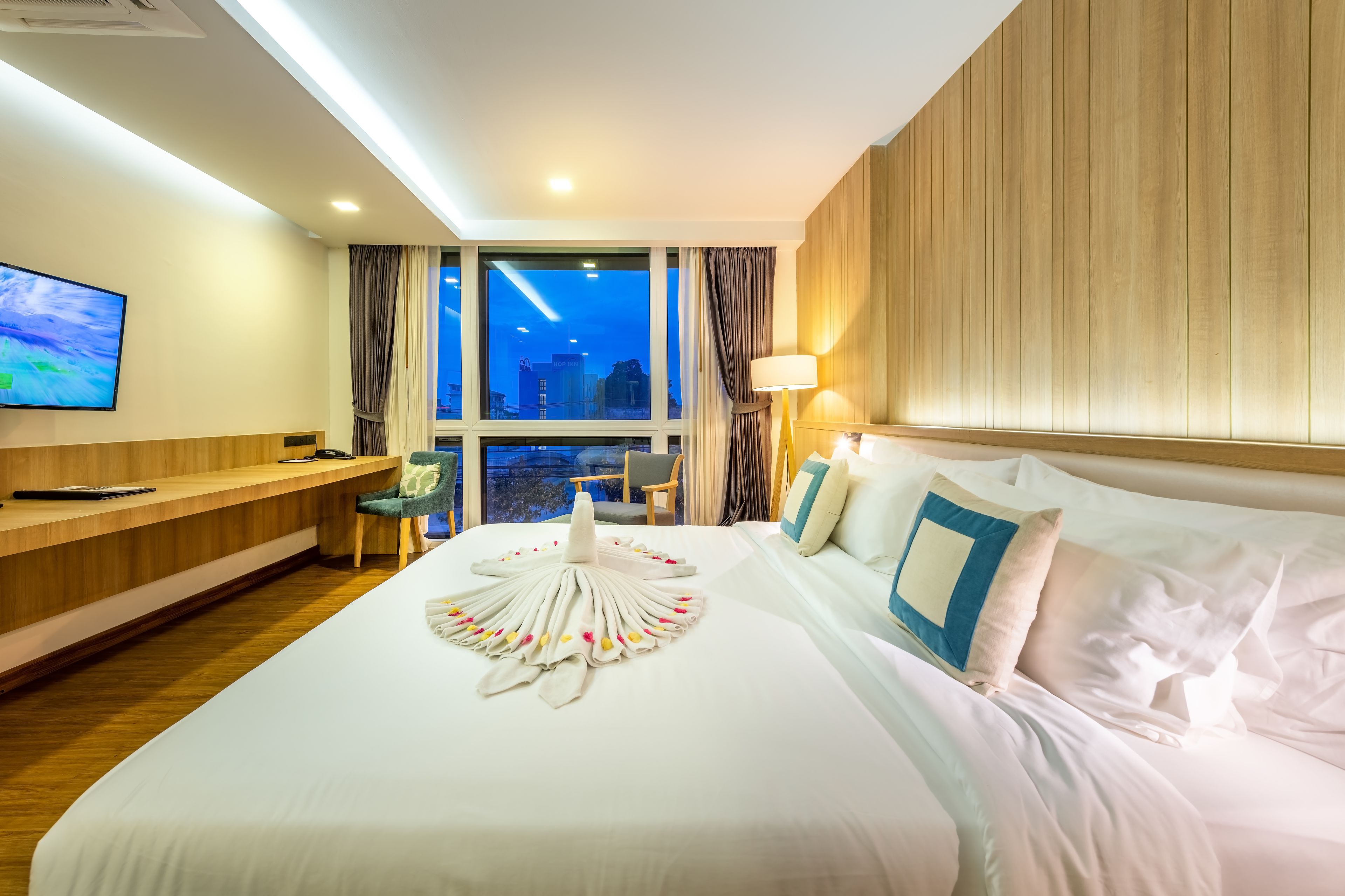 Habitación de hotel con una cama grande, un televisor a la izquierda, un escritorio y vistas de la ciudad al atardecer.