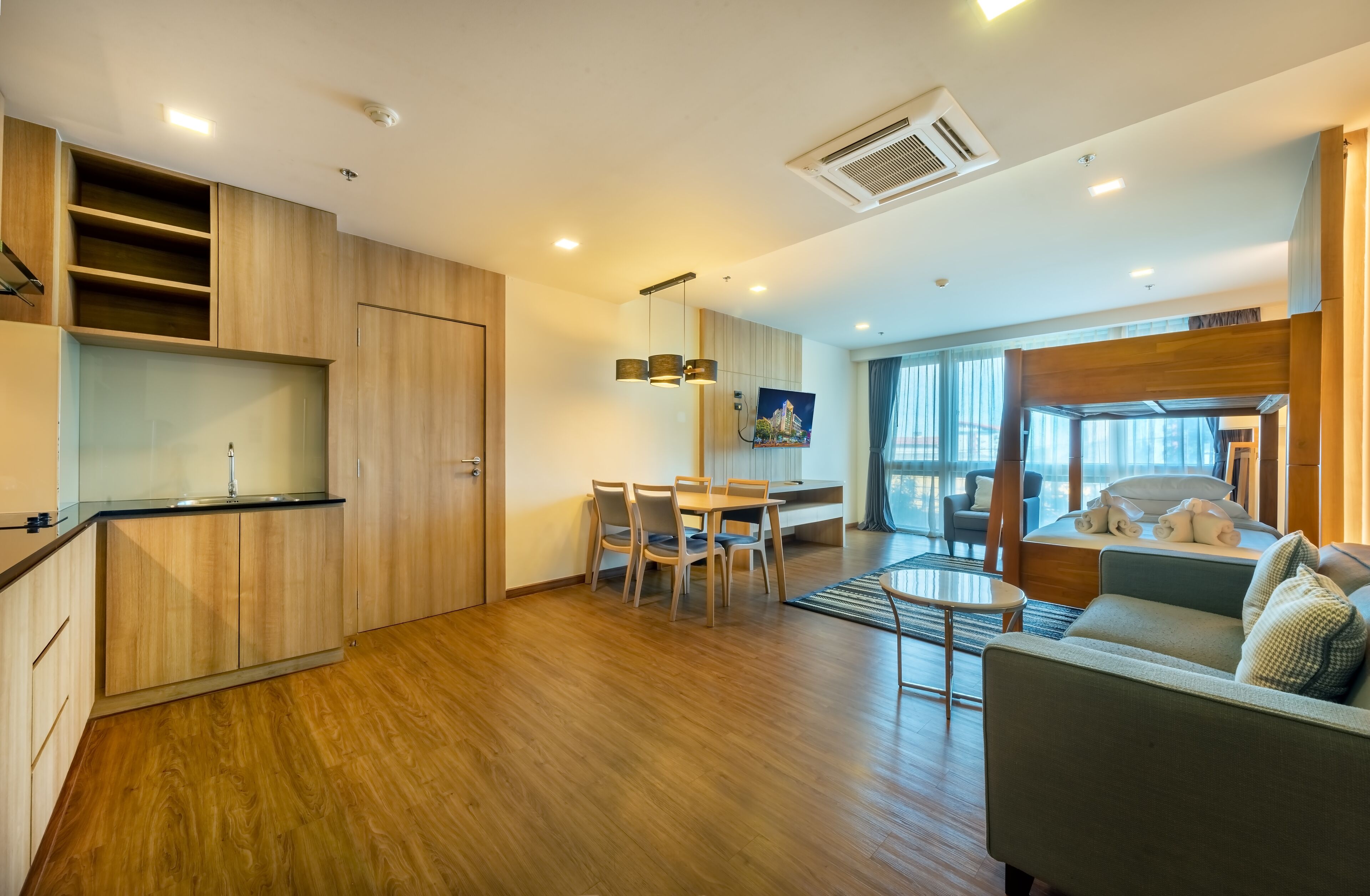 A modern hotel room with a wooden floor, a dining area with a table and chairs, a sofa, and a large window providing a view.