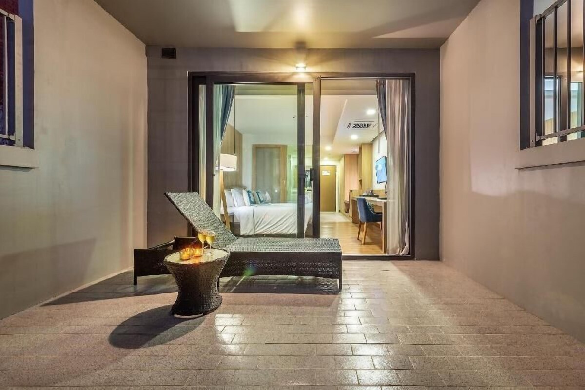 A modern hotel room with a bed, a small table with a drink, and a seating area with a fire feature.
