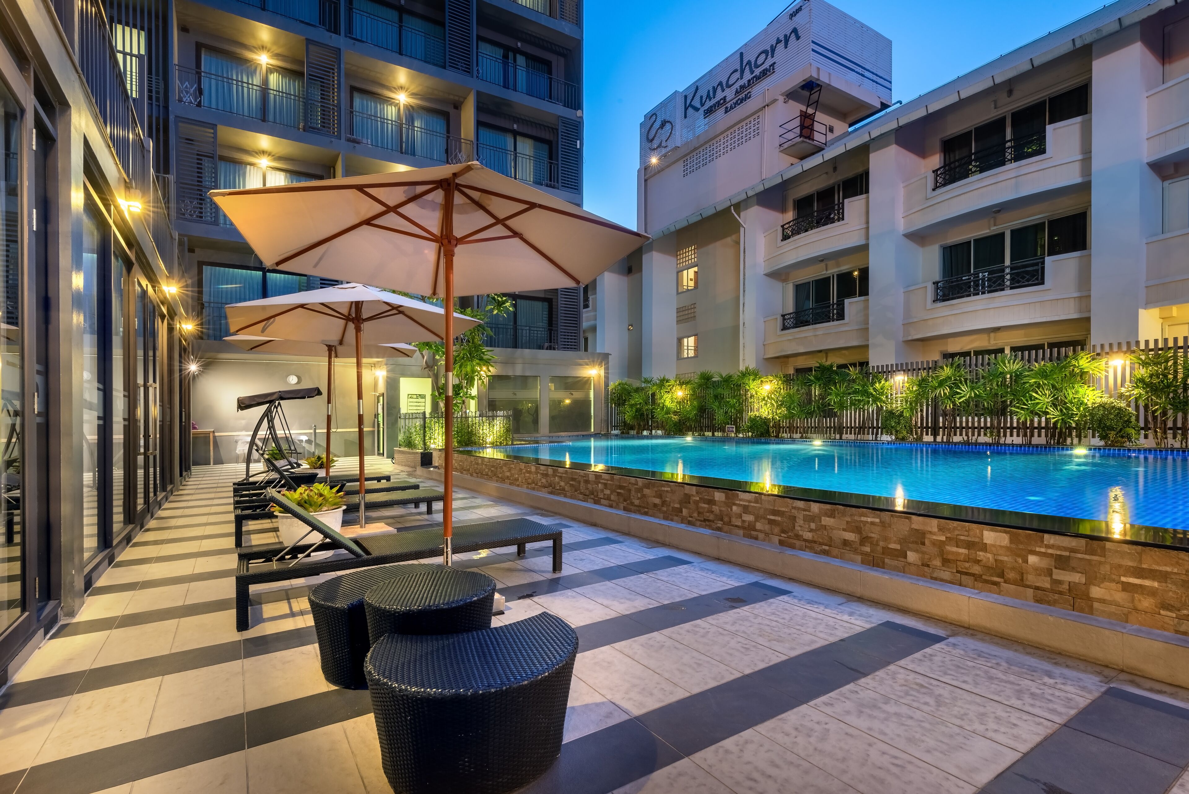 Un área de piscina en la azotea con sillones de descanso y sombrillas, rodeada por un edificio moderno con balcones y un letrero que dice 'Kunchorn Apartment Hotel'.