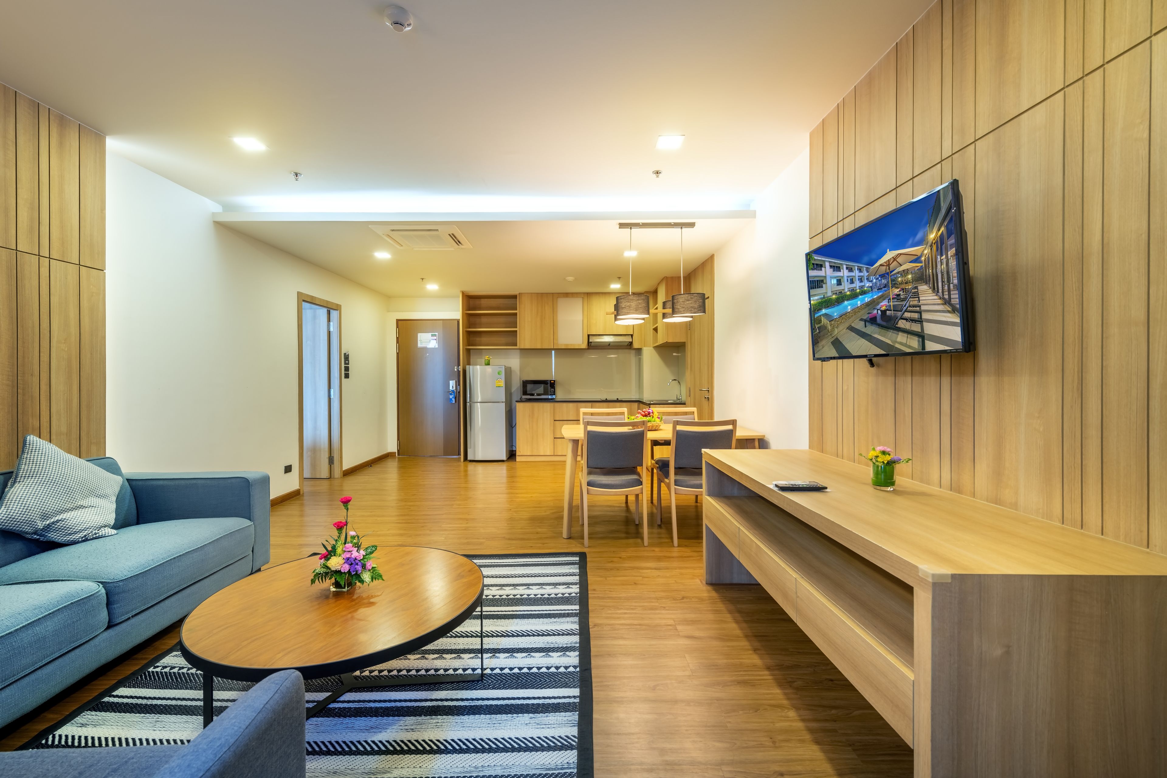 A modern living room with a sofa, coffee table, and a dining area with chairs and a table. A TV is mounted on the wall, and there is a kitchen in the background.