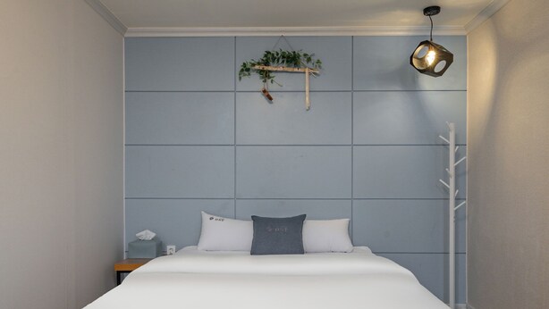 A neatly made bed with white and gray bedding, a bedside table with a tissue box, and a hanging plant on the wall above the bed. - OST Hotel (Chuncheon)