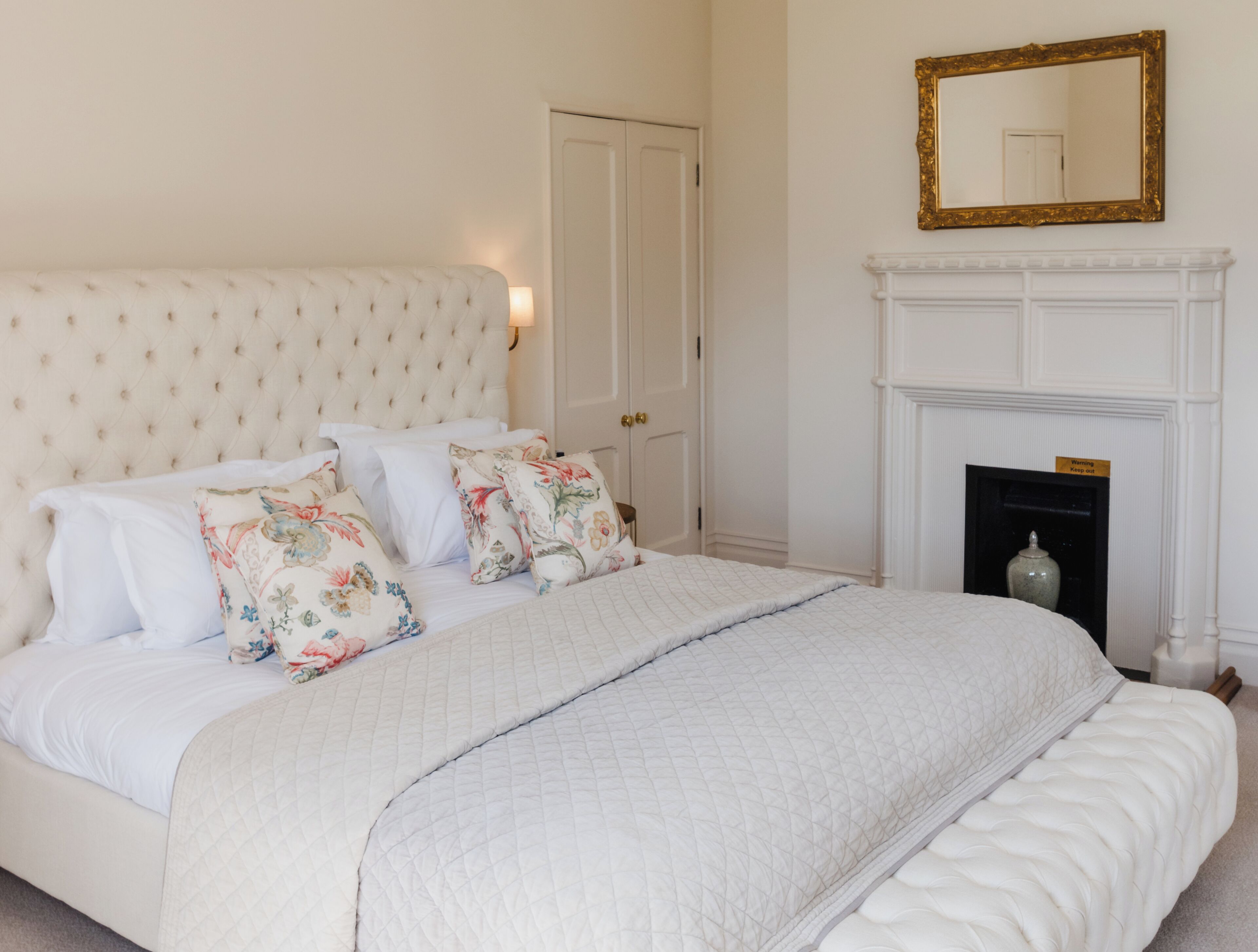 Een slaapkamer met een groot bed, bloemenkussens, een geborduurde sprei, een witte deur, een open haard en een spiegel aan de muur.