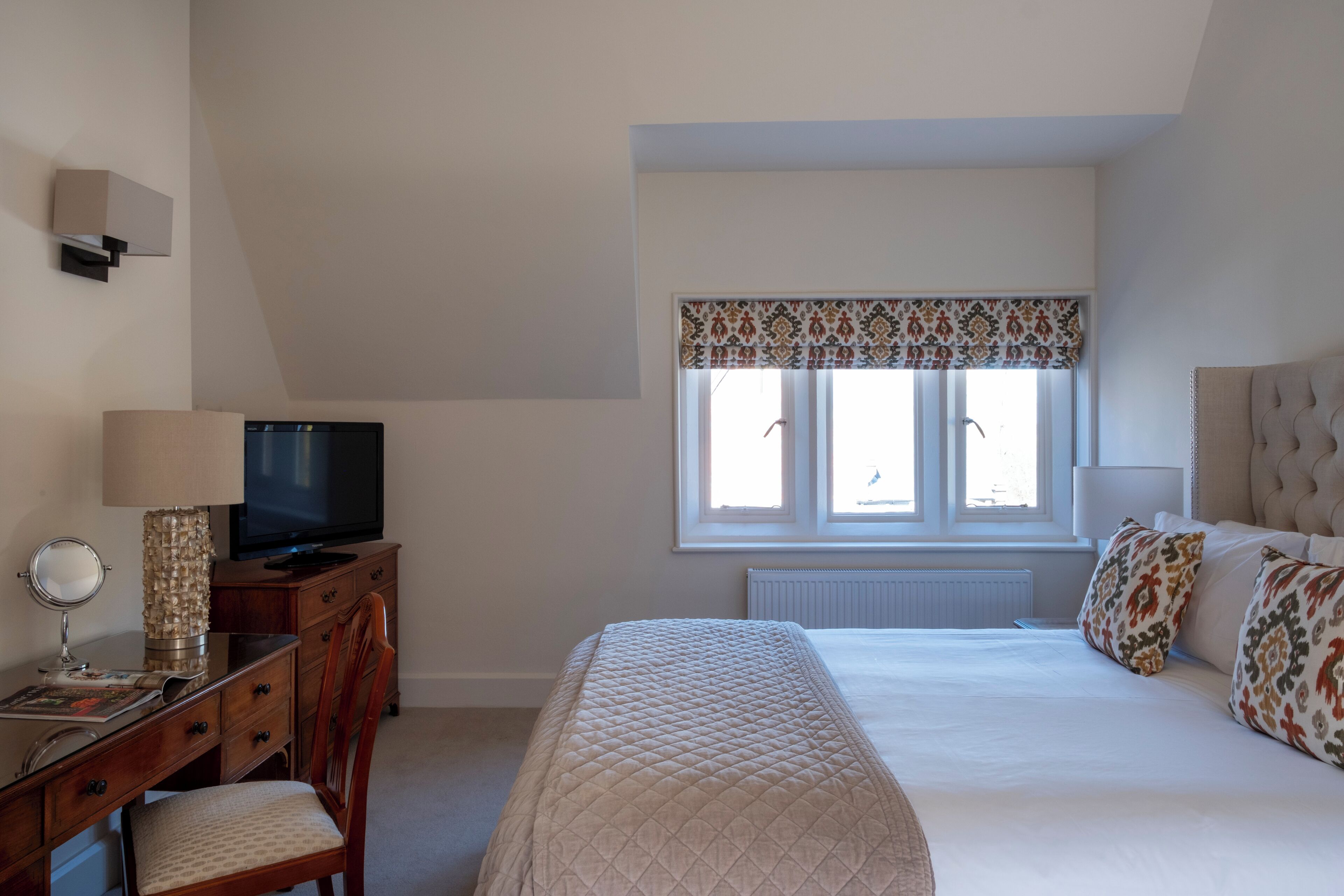 A bedroom with a bed, a desk with a television, a chair, and a window with patterned curtains.