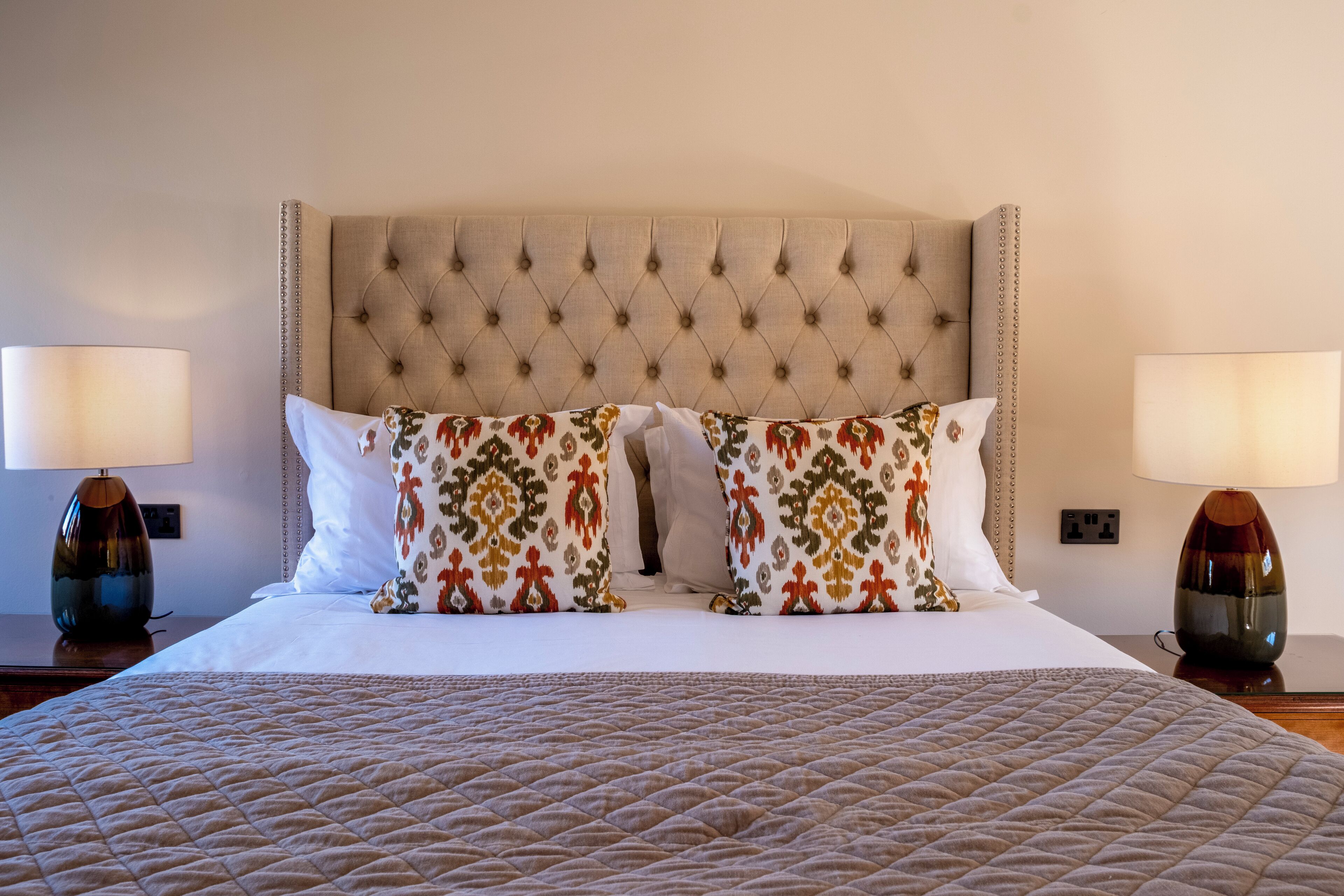 A neatly made bed with a quilted cover, a tufted headboard, and two matching lamps on either side.