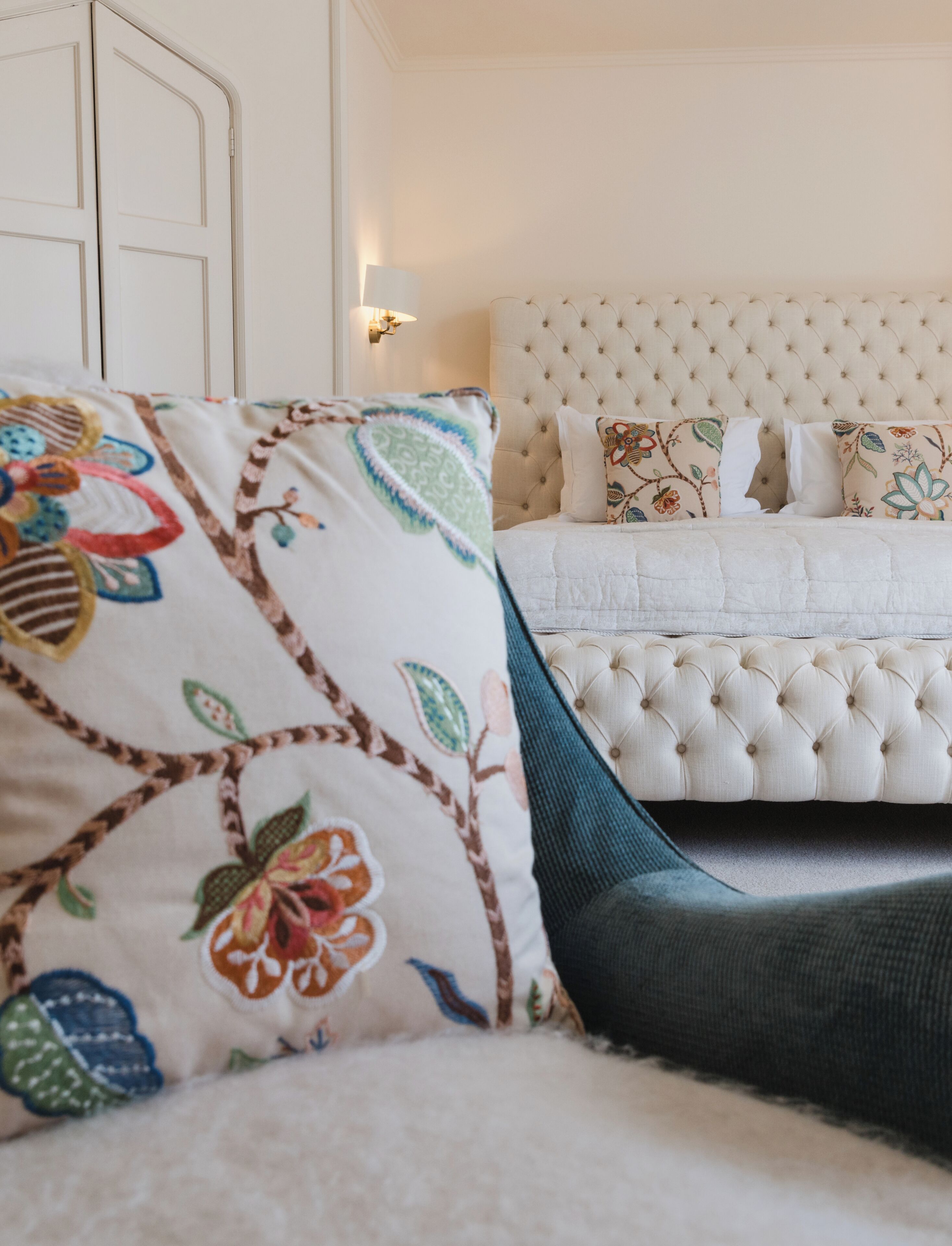 Un dormitorio con cabecera capitonada, almohadas estampadas con motivos florales y una cama con edredón blanco.