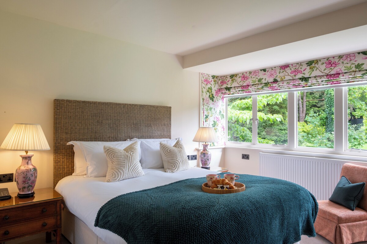 Un dormitorio con una cama grande, cabecera de mimbre, ventana con cortinas florales y una mesita de noche con lámpara y un florero.