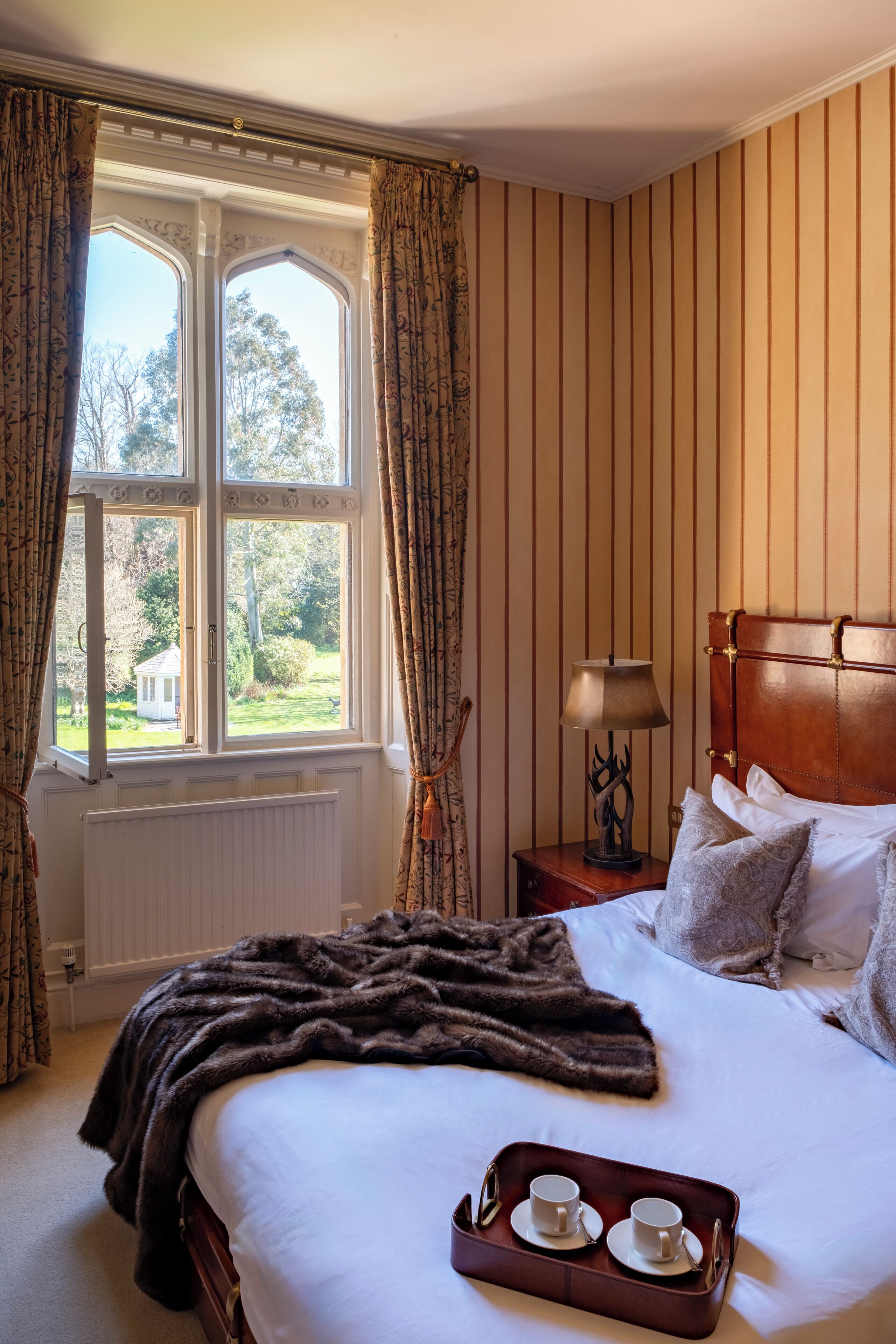 Habitación de hotel con una cama grande, cabecera de madera, ventana con cortinas, mesita de noche con lámpara y bandeja con tazas y platillos.