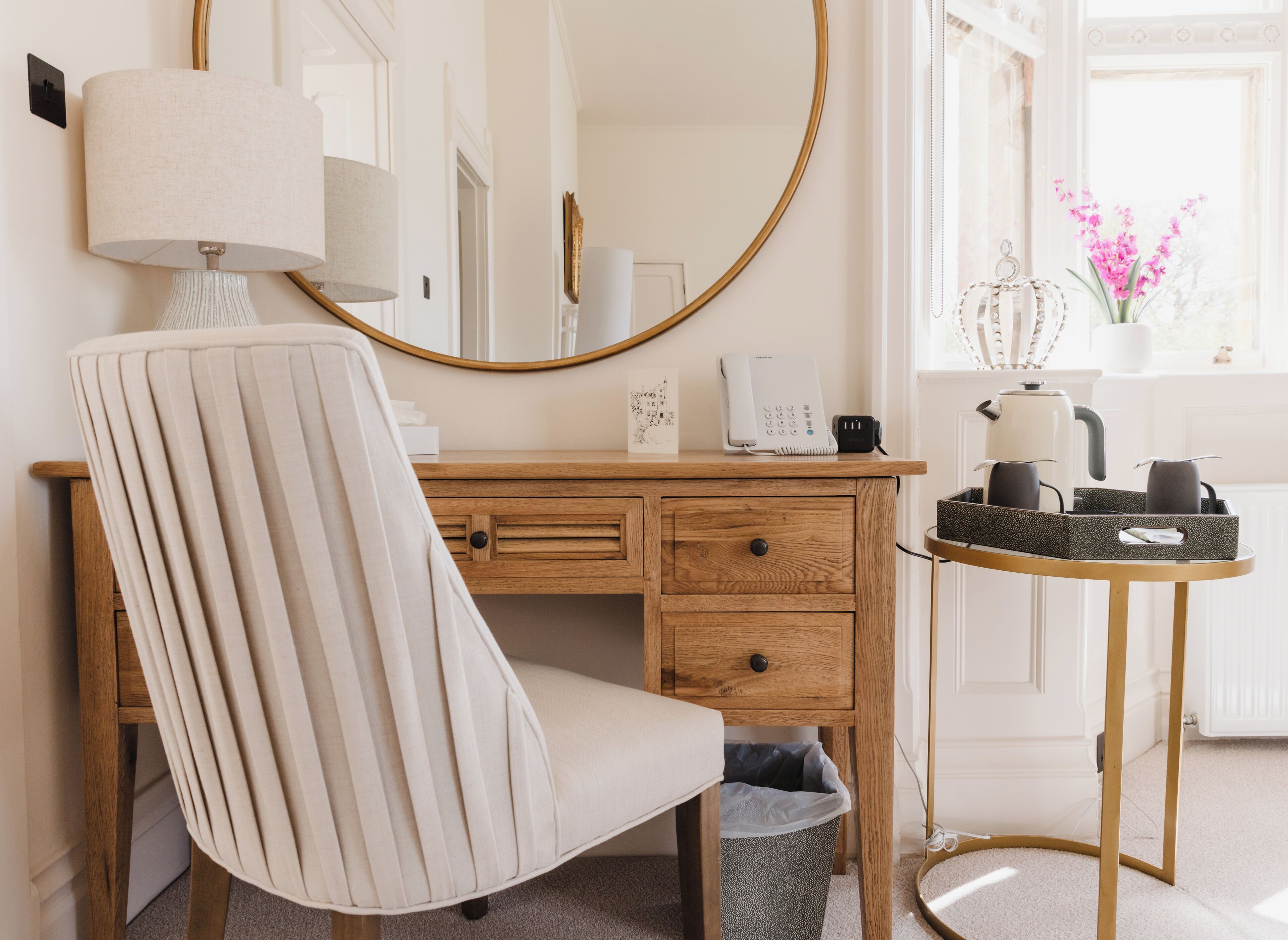 A room with a wooden desk, a chair, a mirror, a lamp, a telephone, a coffee maker, and a small table with a tray.