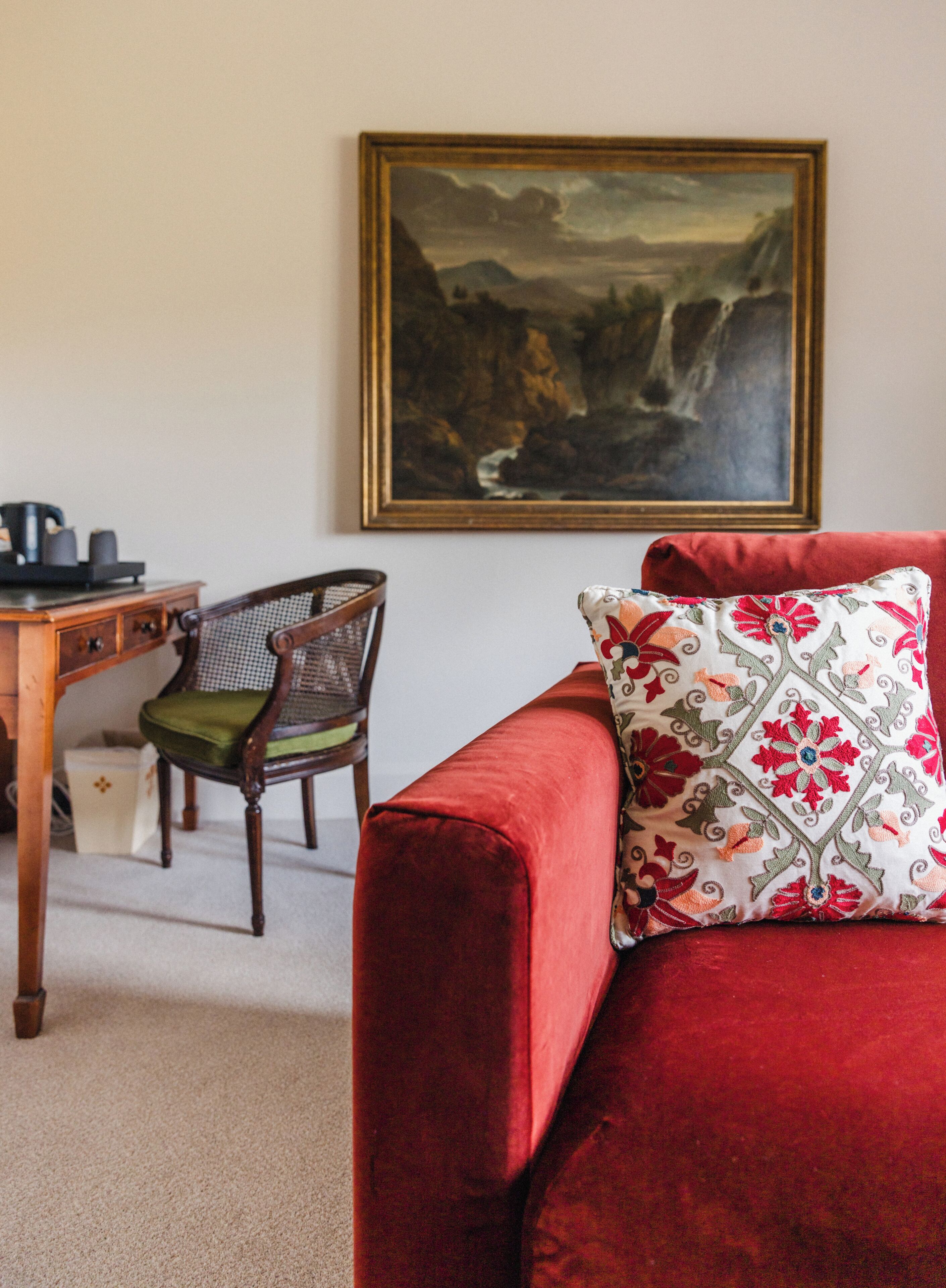 A red sofa with a floral pillow, a wooden chair with a green cushion, a desk with a coffee maker, and a framed painting on the wall.