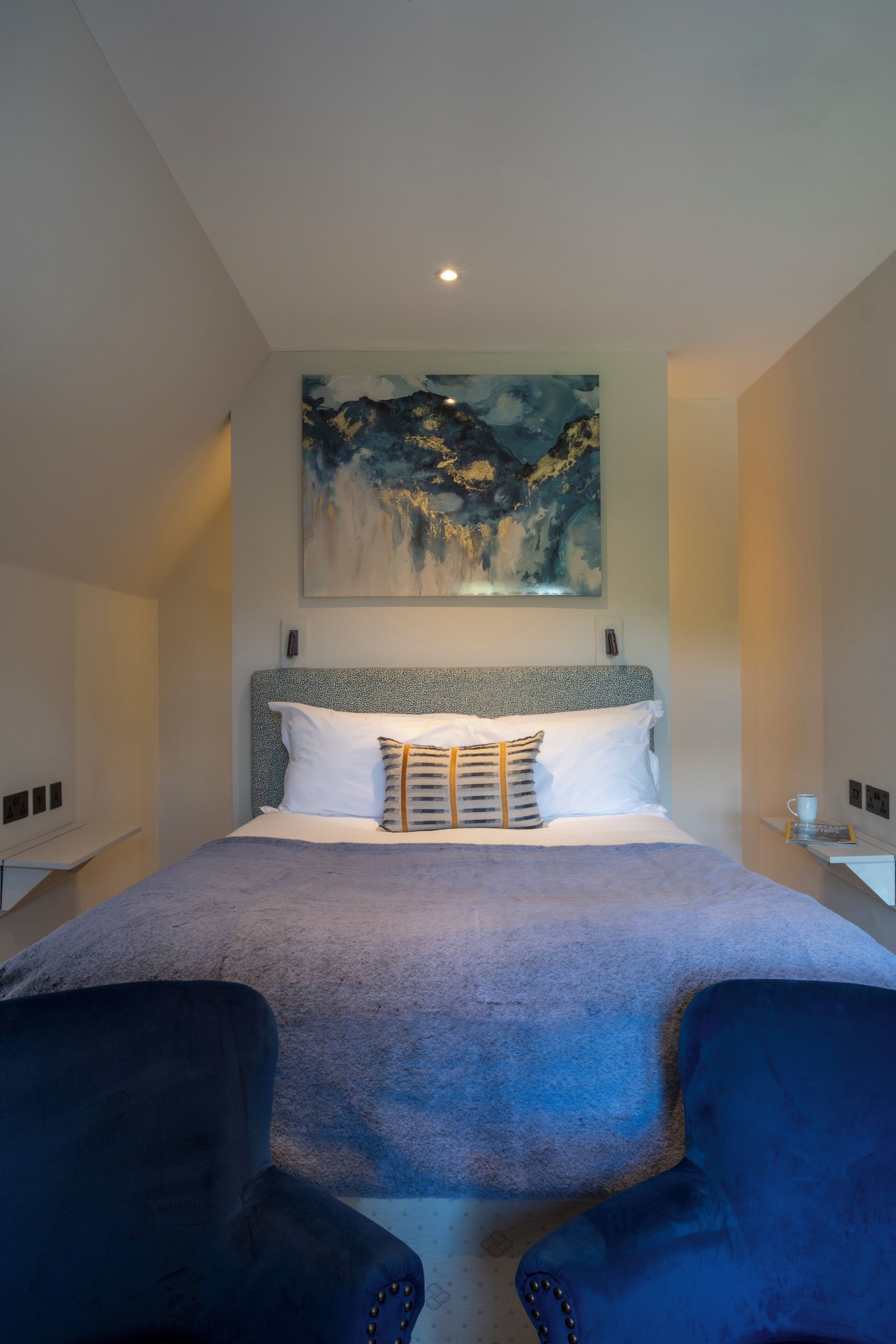 A modern hotel room with a large bed, a painting above it, and two armchairs.