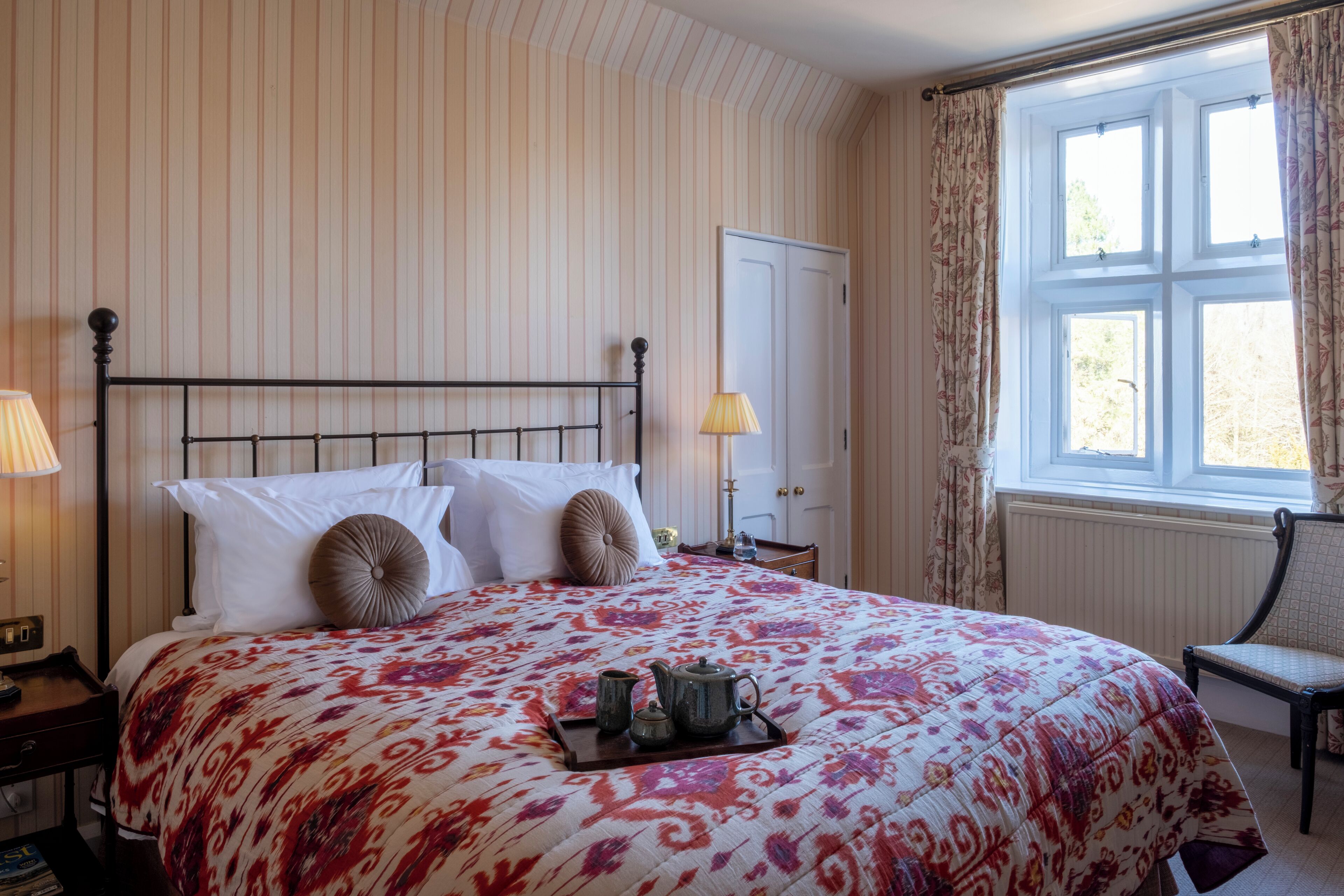 A neatly made bed with a floral bedspread, a tray with a teapot and cups, and a chair by a window with curtains.