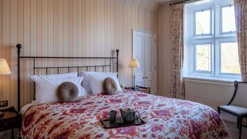 A neatly made bed with a floral bedspread, a tray with a teapot and cups, and a chair by a window with curtains.