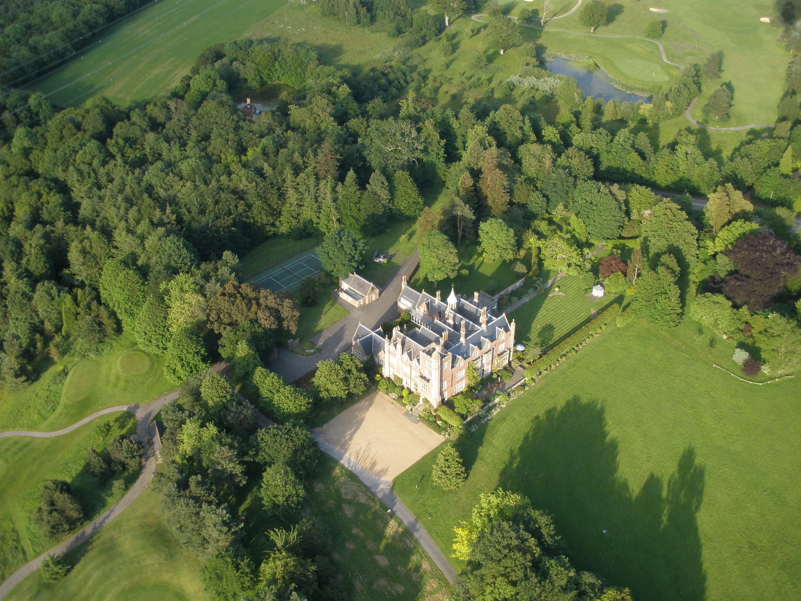 An aerial view of a large estate with a main building, tennis court, and surrounding green fields.