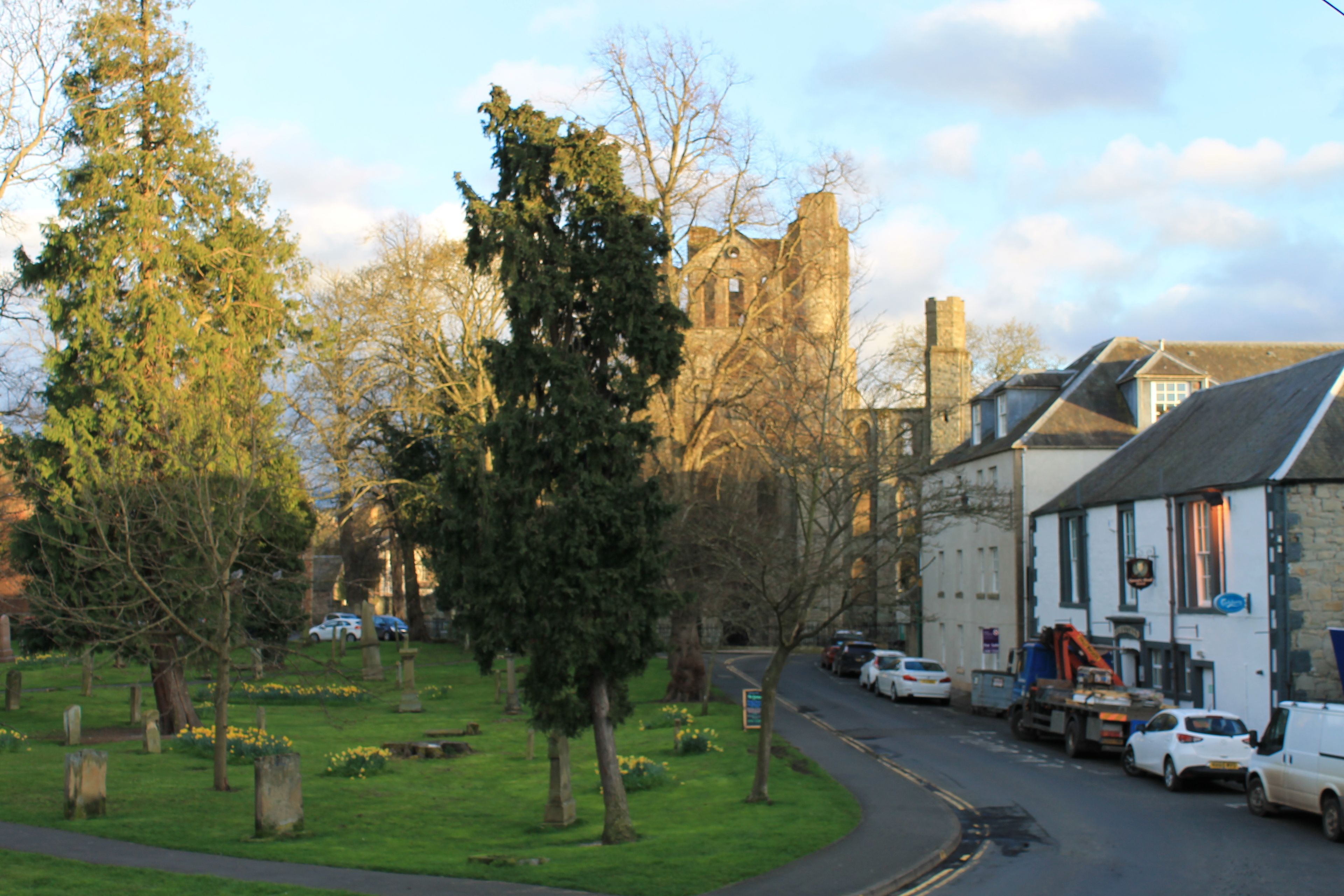 A street view with a historic building, parked cars, and a grassy area with trees.