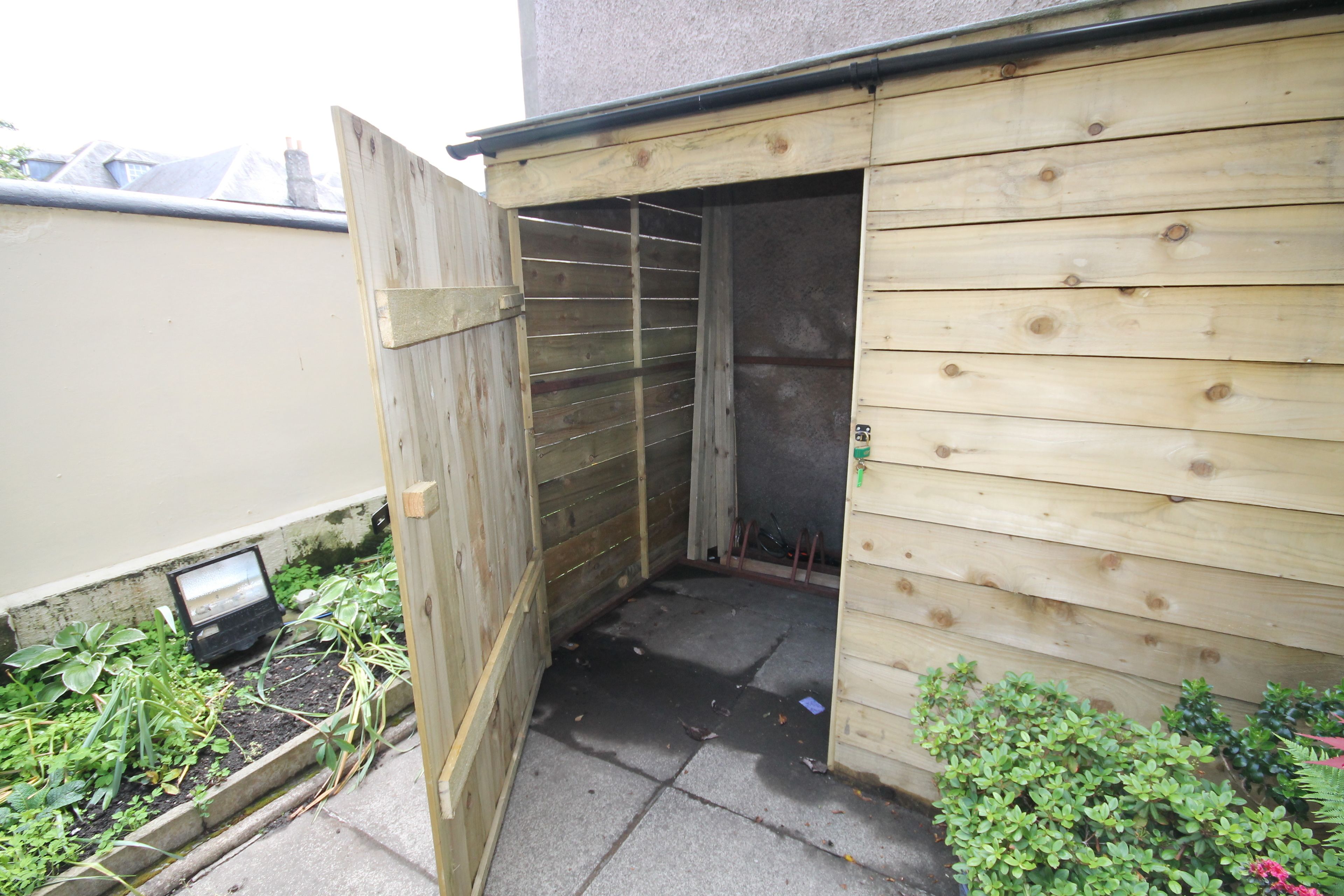 A wooden shed with a metal door, a small garden area with plants, and a concrete floor.