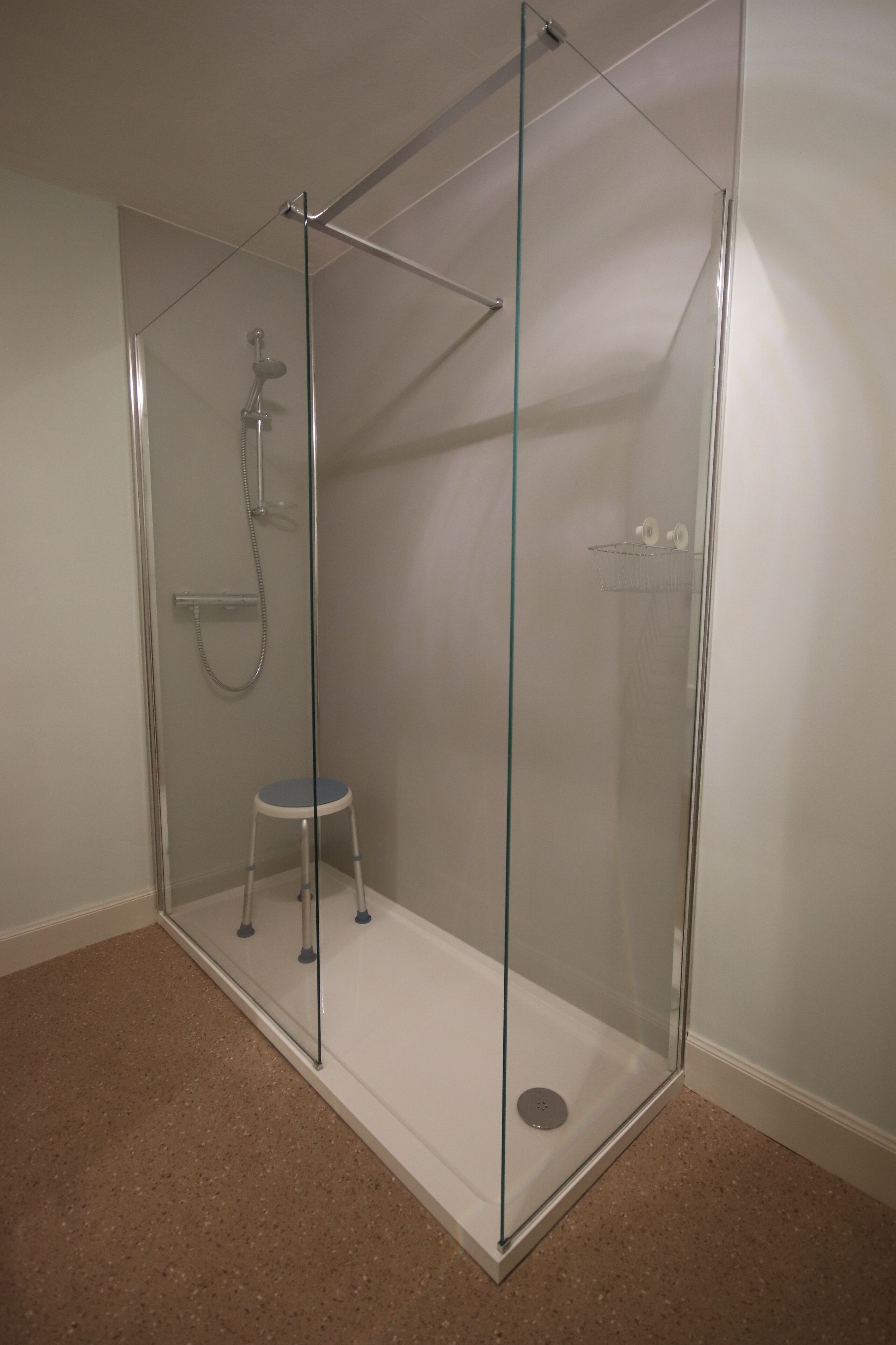 Un espace douche fermé par des parois de verre, comprenant un pommeau de douche, un petit tabouret et une porte en verre.
