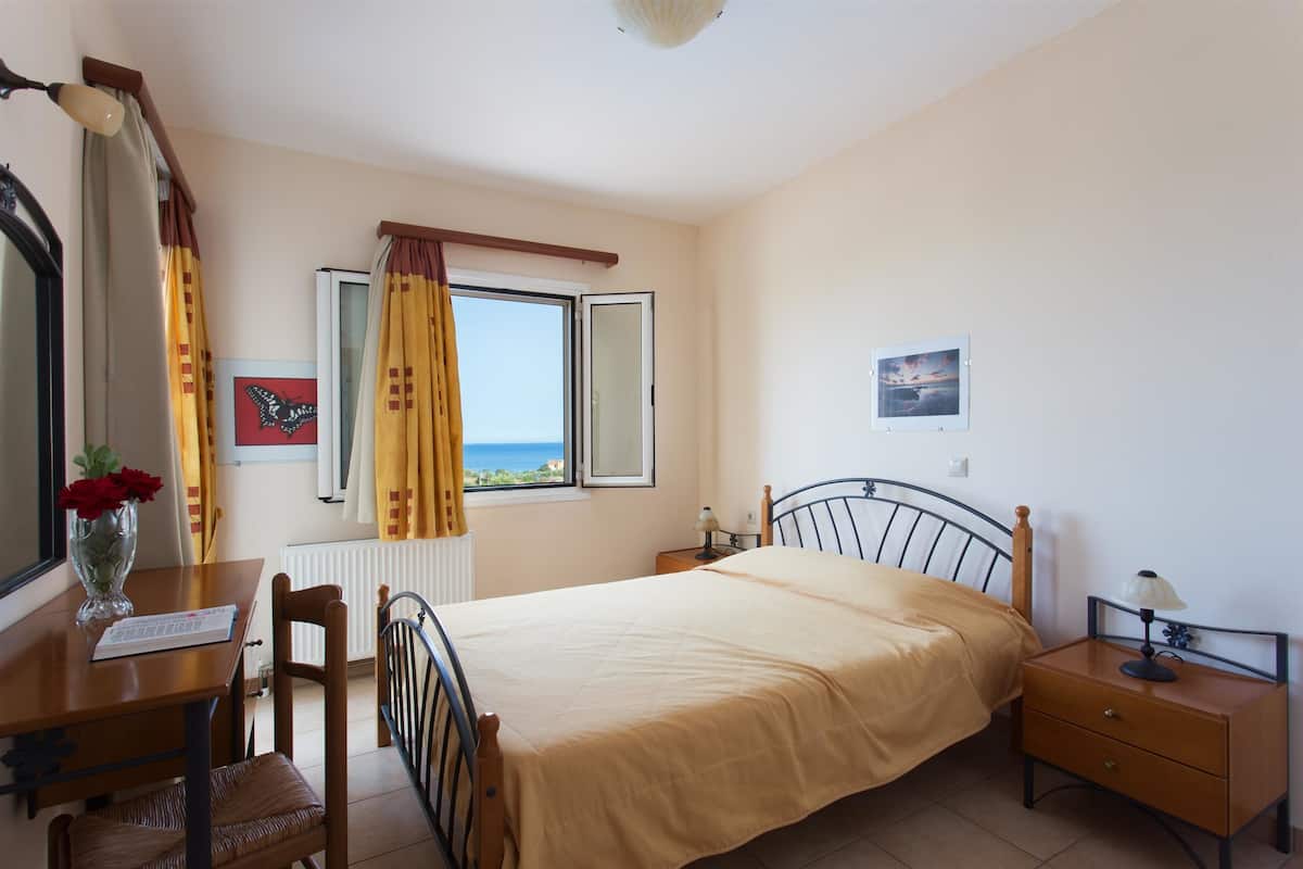 Ein Schlafzimmer mit Bett, Schreibtisch, Stuhl, Nachttisch, Lampe, einem Fenster mit Vorhängen und einem Bild an der Wand.