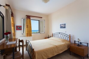 Un dormitorio con cama, escritorio, silla, mesita de noche, lámpara, ventana con cortinas e imagen en la pared.