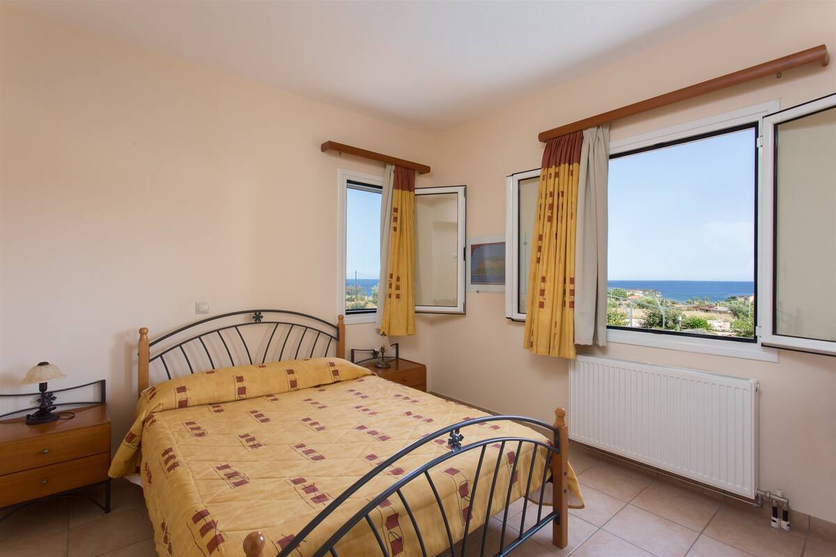 Ein Schlafzimmer mit Bett, Nachttisch, Fenster mit Vorhängen und Meerblick.