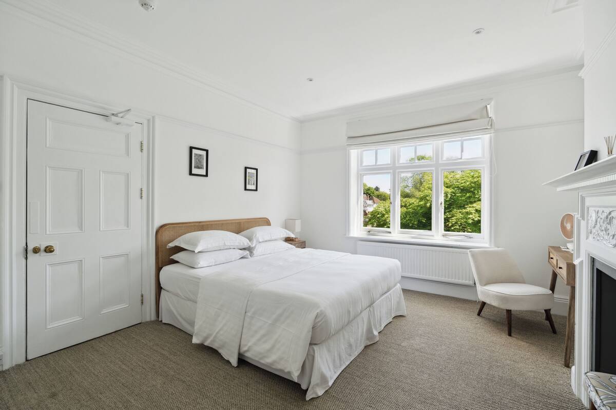 A bedroom with a large bed, a window with a view of greenery, a chair, and a fireplace.