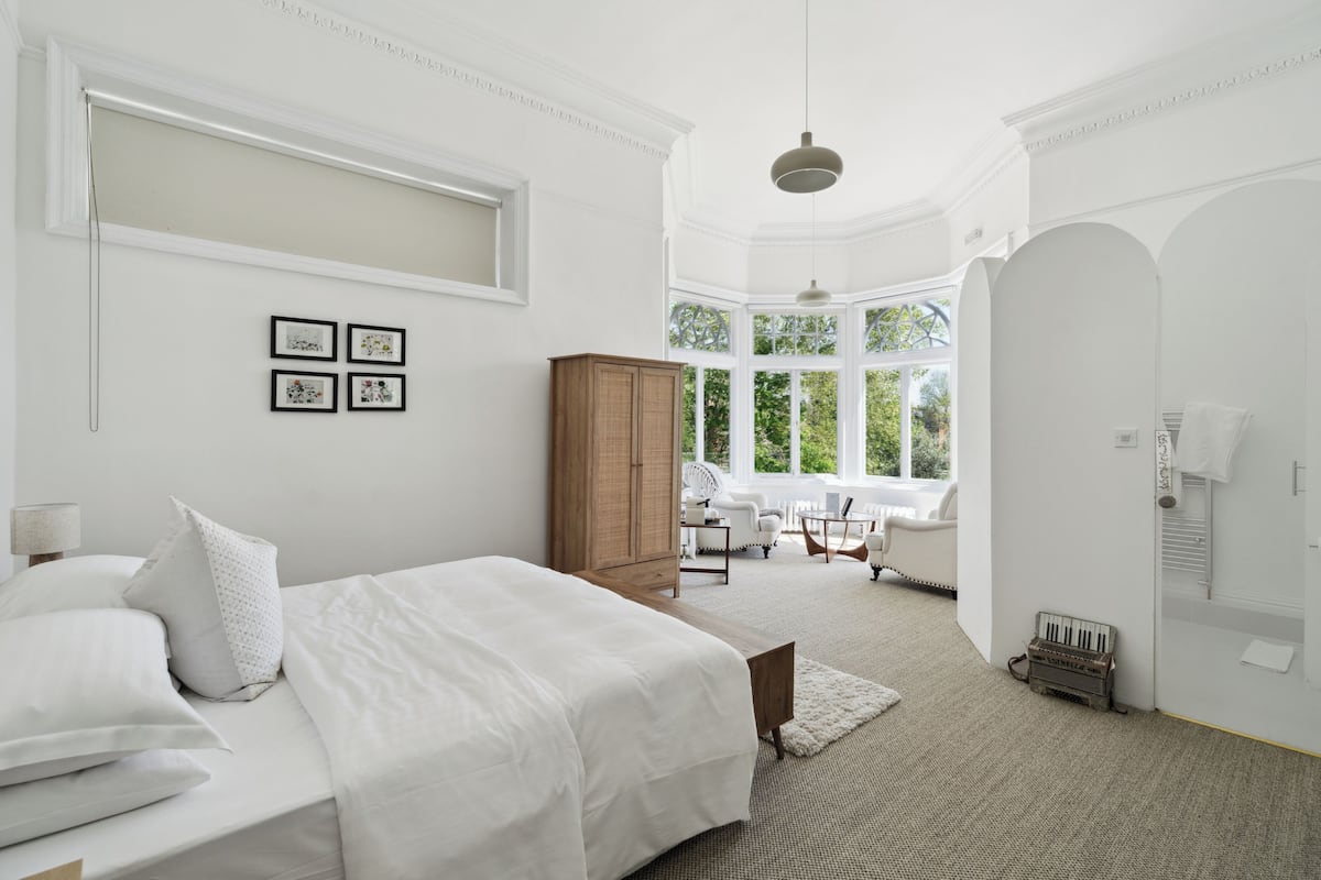 A spacious bedroom with a large bed, a wooden wardrobe, a sitting area with a table and chairs, and a view of greenery through large windows.