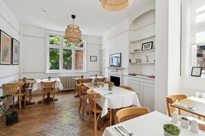 A dining area with wooden tables and chairs, white walls, and a large window with a view of greenery. - River Arts Club (Maidenhead)