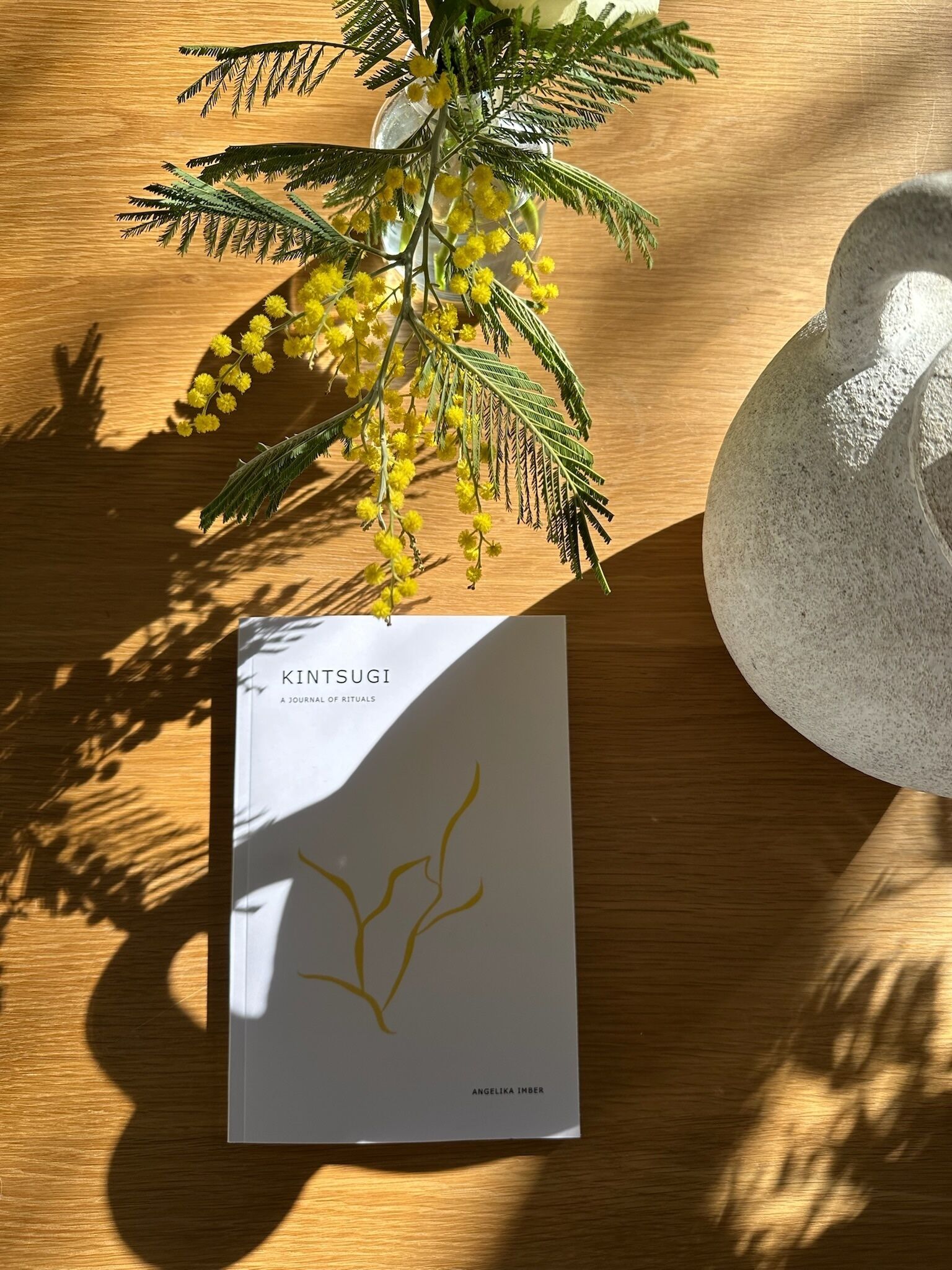 A book titled "KITSUGI A JOURNAL OF RITUALS" by Angelika Imber on a wooden surface with a decorative plant and a stone sculpture.