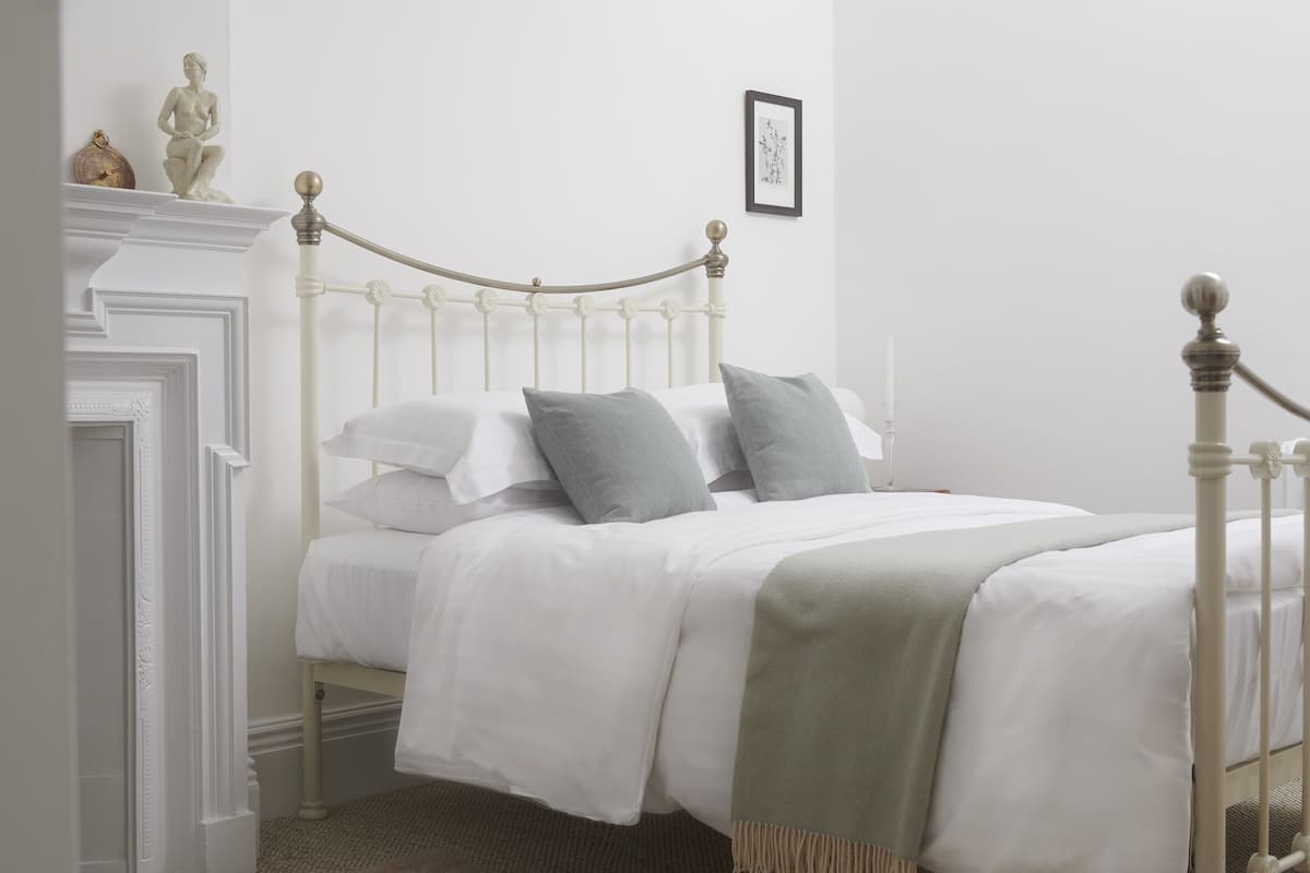 A neatly made bed with white bedding and a light green throw blanket, a white metal bed frame, and a small statue on a mantelpiece.