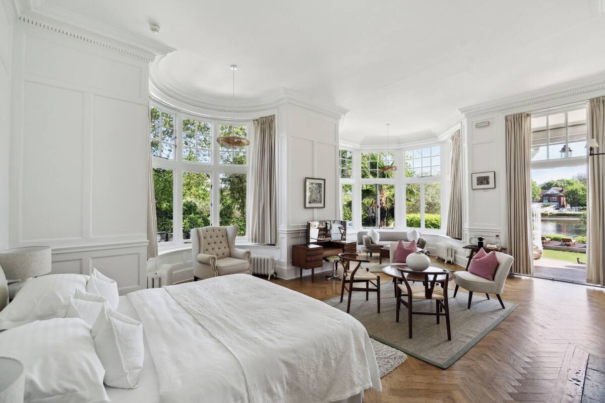 A spacious bedroom with a large bed, a desk, and a seating area by a window overlooking a garden.