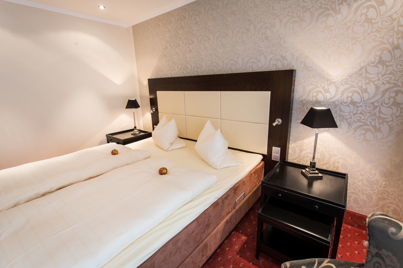 Ein Hotelzimmer mit zwei Einzelbetten, einem Holz-Kopfteil, einem Nachttisch mit Lampe und einem roten Teppichboden.