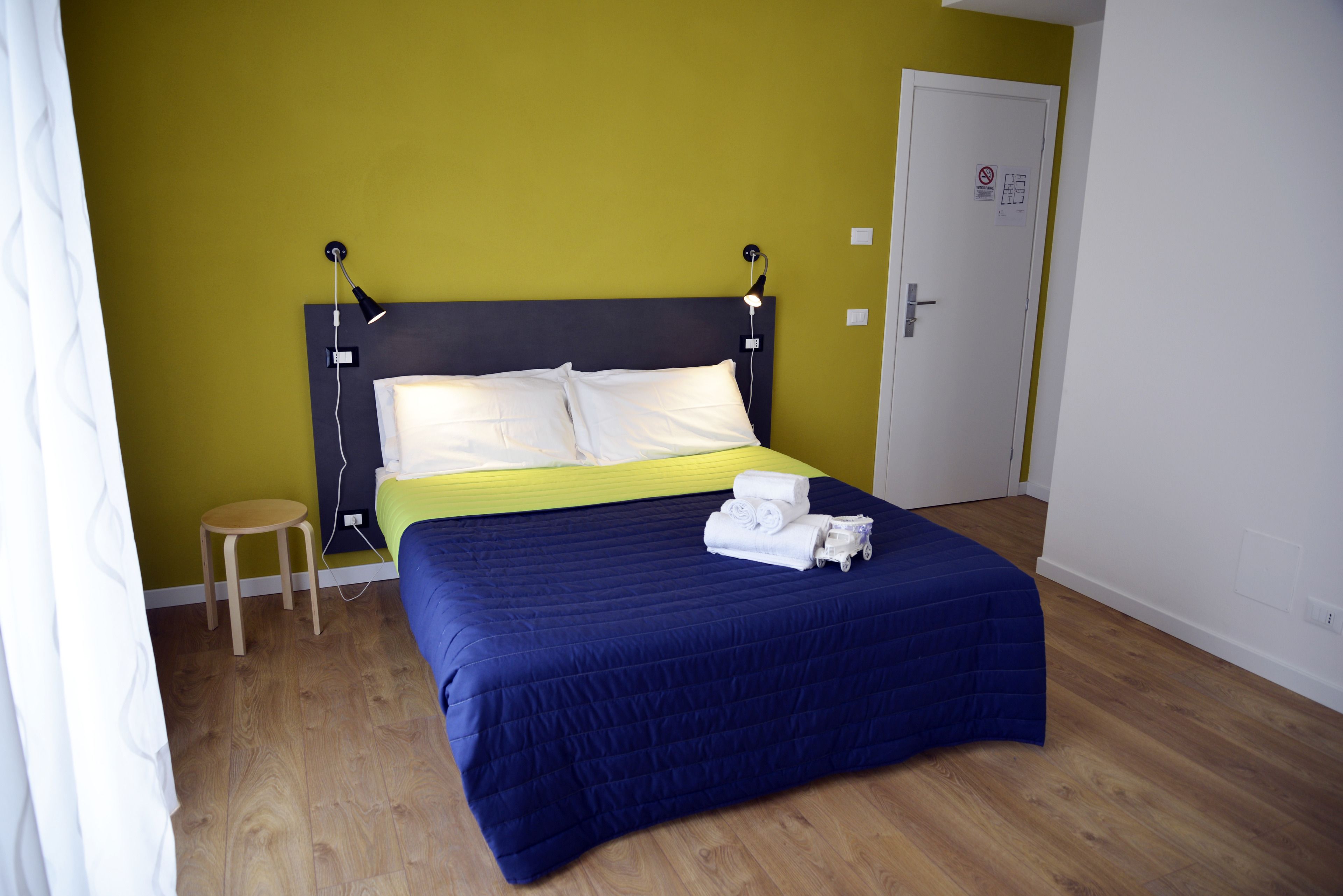 Una cama bien tendida con una colcha azul y almohadas blancas, un taburete de madera pequeño y una puerta con un cartel.