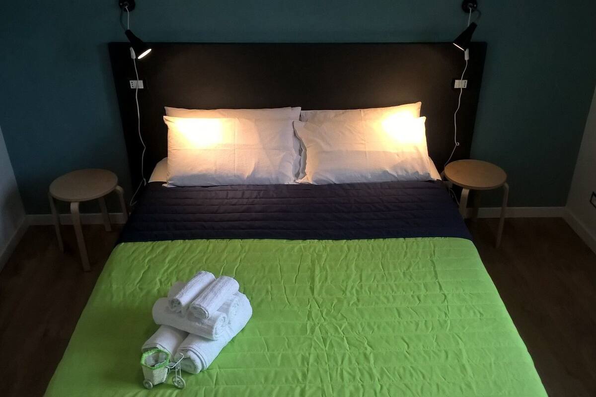 A neatly made bed with a green bedspread, two bedside lamps, and a stack of rolled towels on the foot of the bed.
