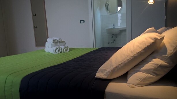Une chambre d’hôtel avec un lit, une literie verte et noire, une pile de serviettes blanches, un miroir et une salle de bain avec un lavabo et un miroir.