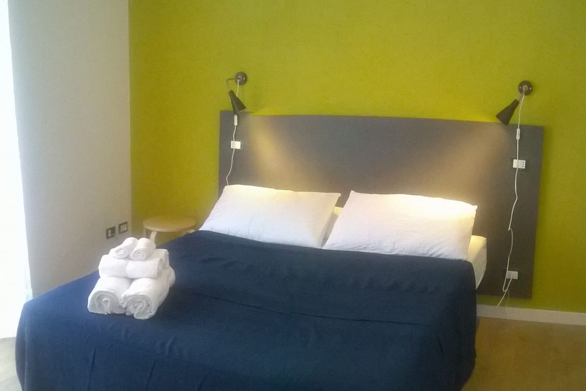 A bed with a dark blue bedspread and two white pillows, a stack of white towels, and a wall-mounted lamp.
