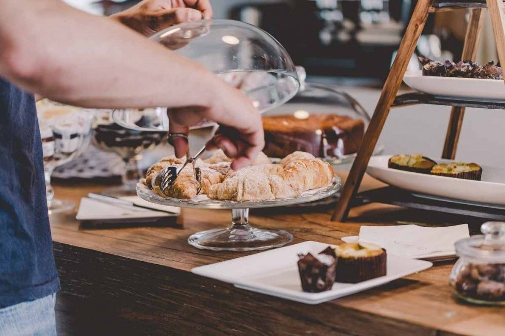 Une personne servant des plats sous un couvercle en verre sur une table de buffet où sont présentés divers pâtisseries et desserts.
