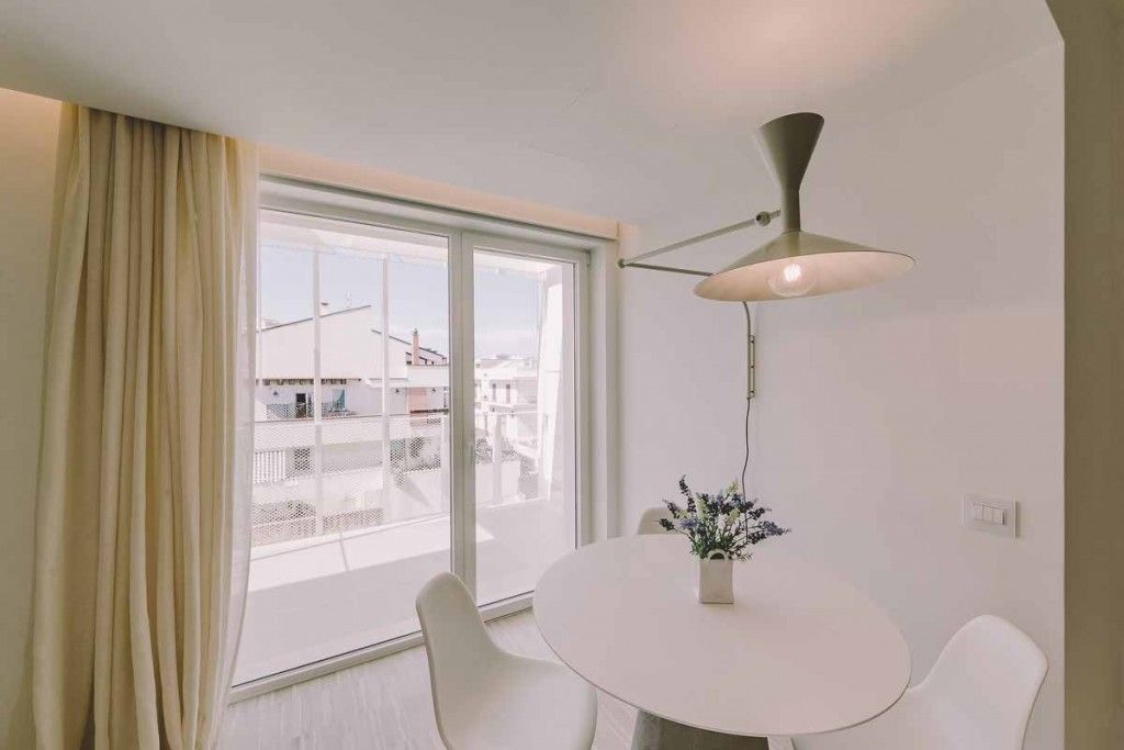 A modern room with a round white table, white chairs, a hanging lamp, and a view of buildings through a large window.