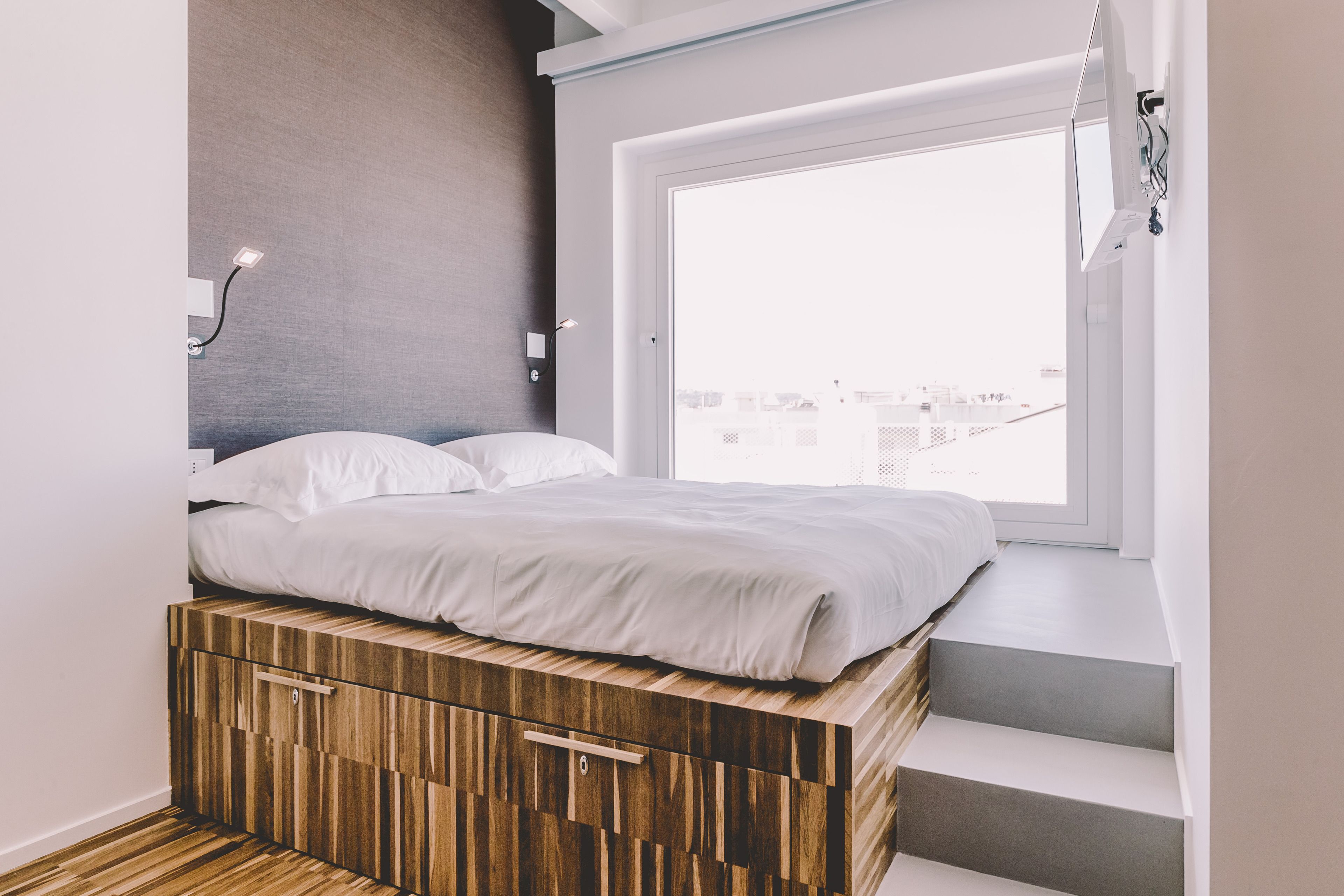A bedroom with a bed, built-in wooden storage, a window with a city view, and a staircase.