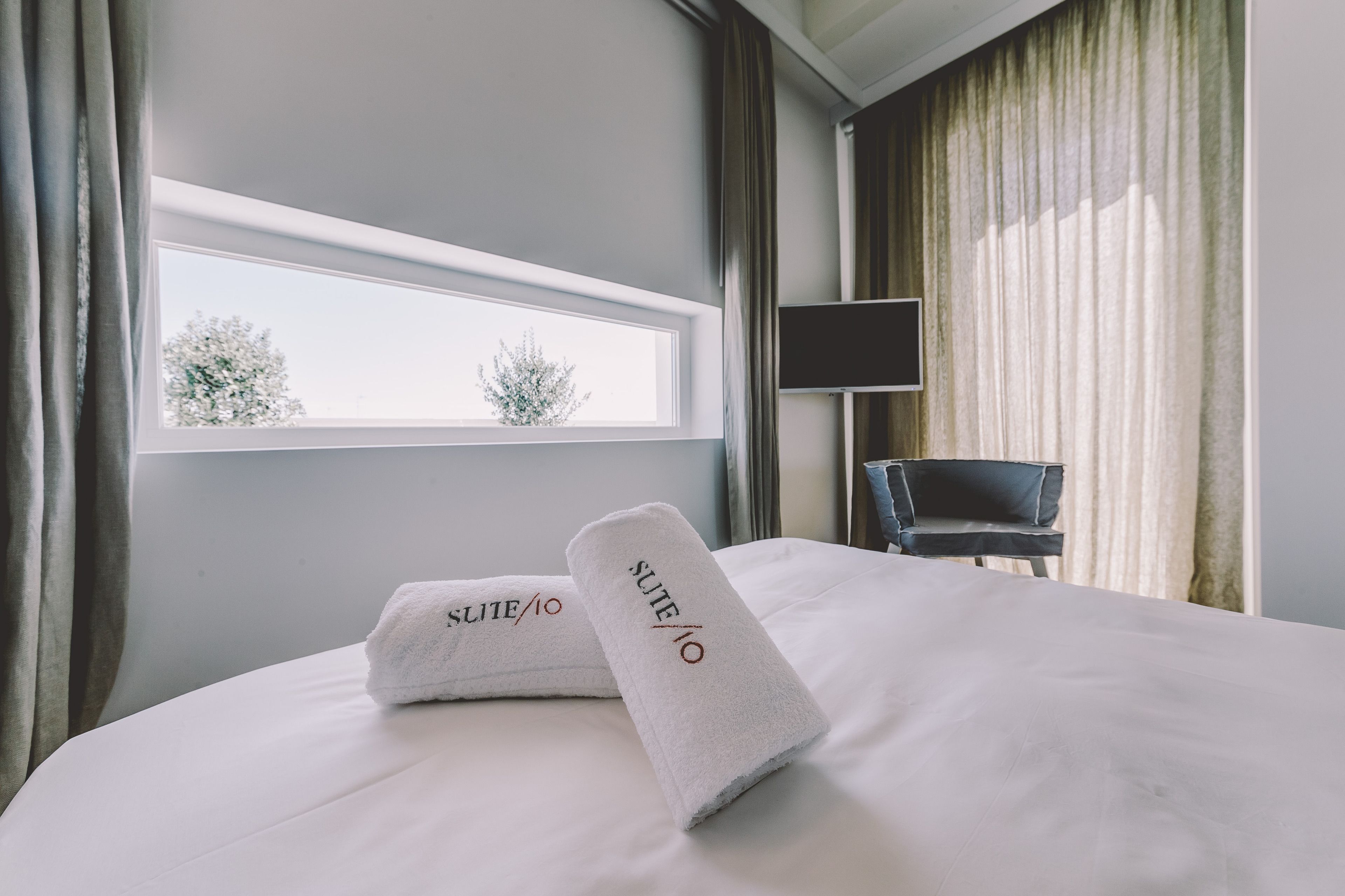 Quarto de hotel com uma janela grande, uma cama com roupa de cama branca, duas toalhas com o texto 'SUITE/10', uma cadeira preta e uma televisão de tela plana.