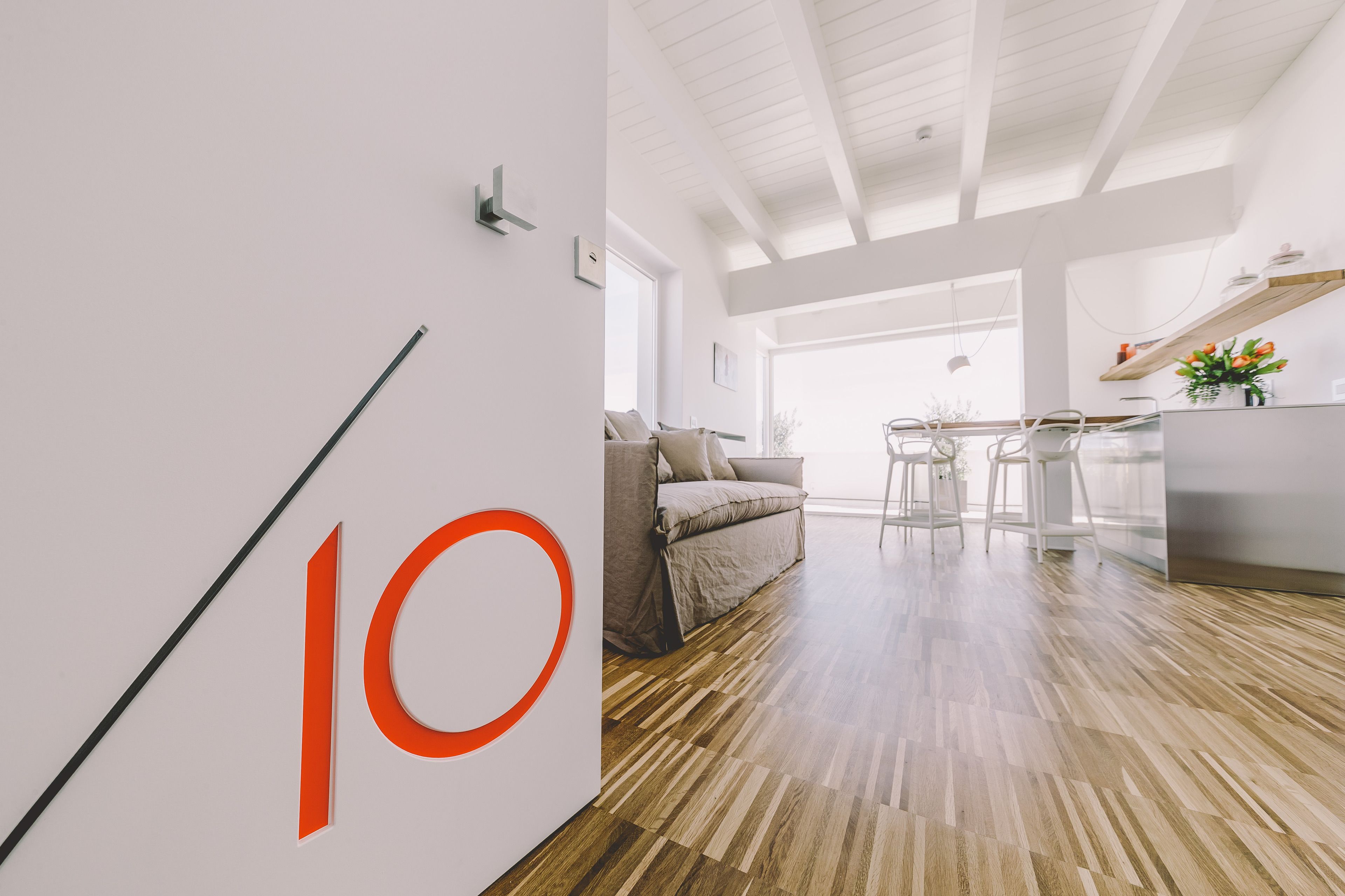 Un intérieur moderne avec un grand chiffre « 10 » peint sur le mur, un plancher en bois, un canapé, un coin repas et une cuisine agrémentée d’une étagère remplie de fleurs.