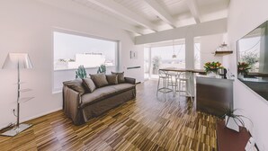 A modern living room with a wooden floor, a sofa, a dining area, and large windows offering a view of the city. - Suite 10 - Home, Design & Spa (Polignano a Mare)
