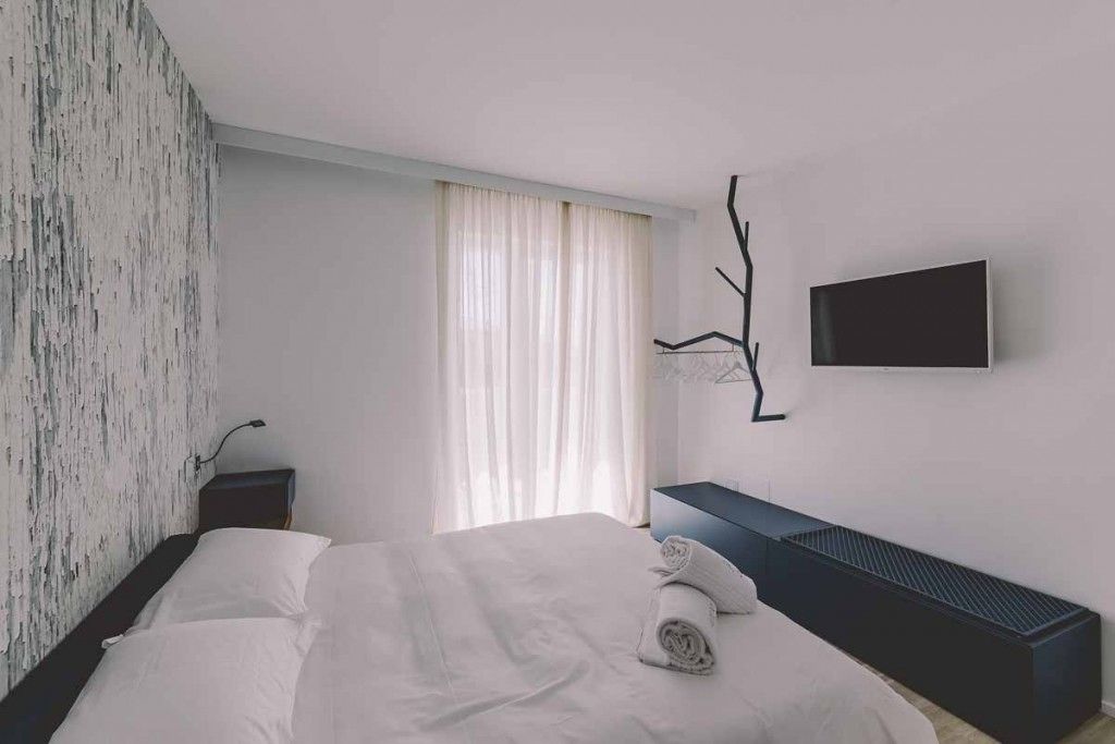 A modern hotel room with a large bed, a flat-screen TV mounted on the wall, and a window with sheer curtains.