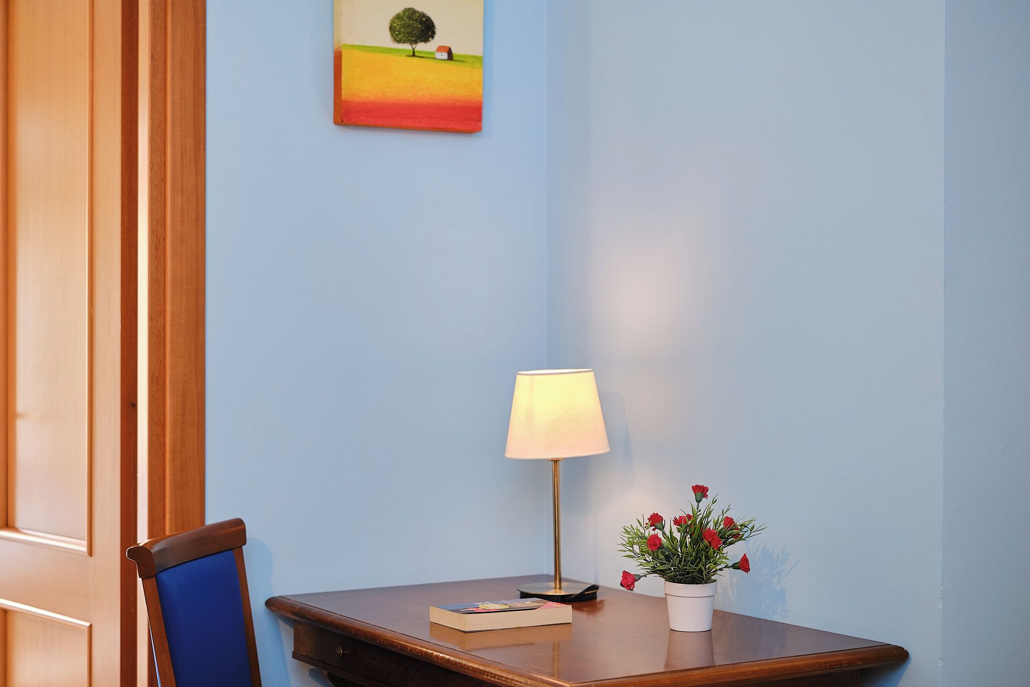 Una mesa de madera con una lámpara, un libro y una maceta con flores rojas.